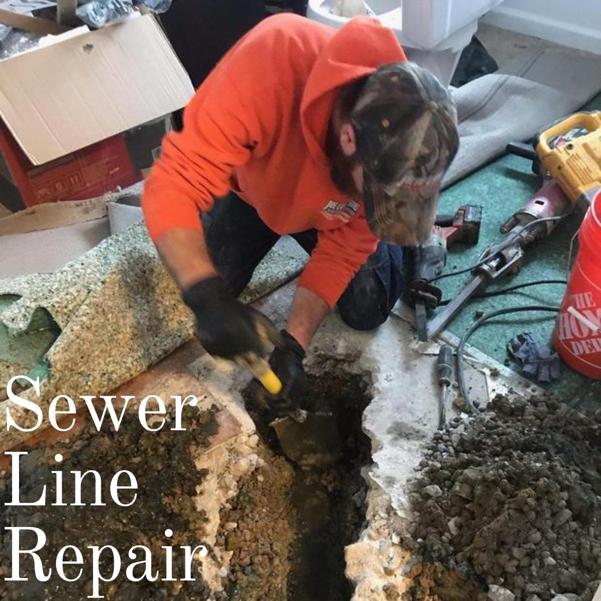 Man in orange hoodie repairs sewer line indoors, using a shovel and tools.