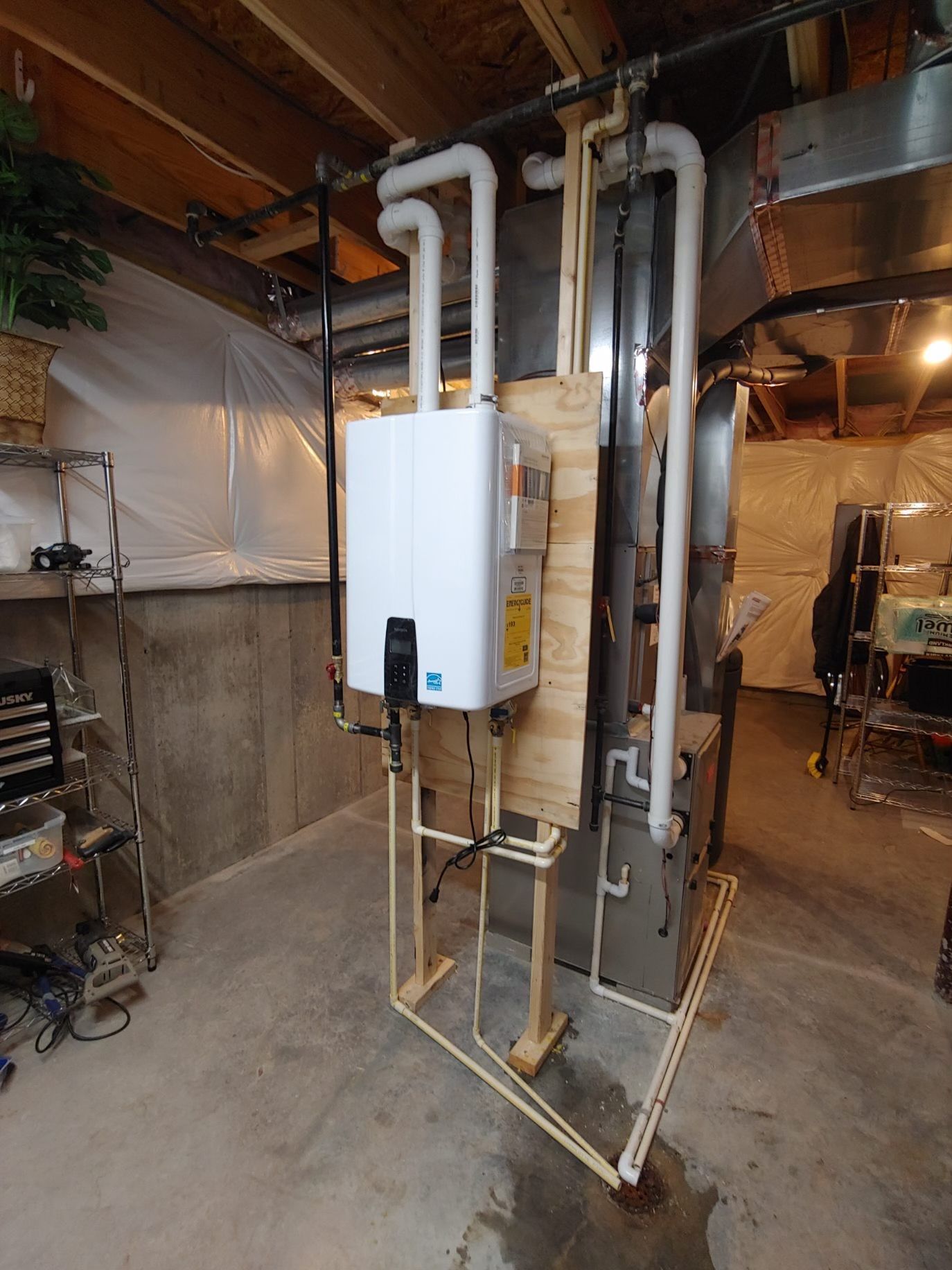 A tankless water heater mounted on plywood in a basement, with associated plumbing.