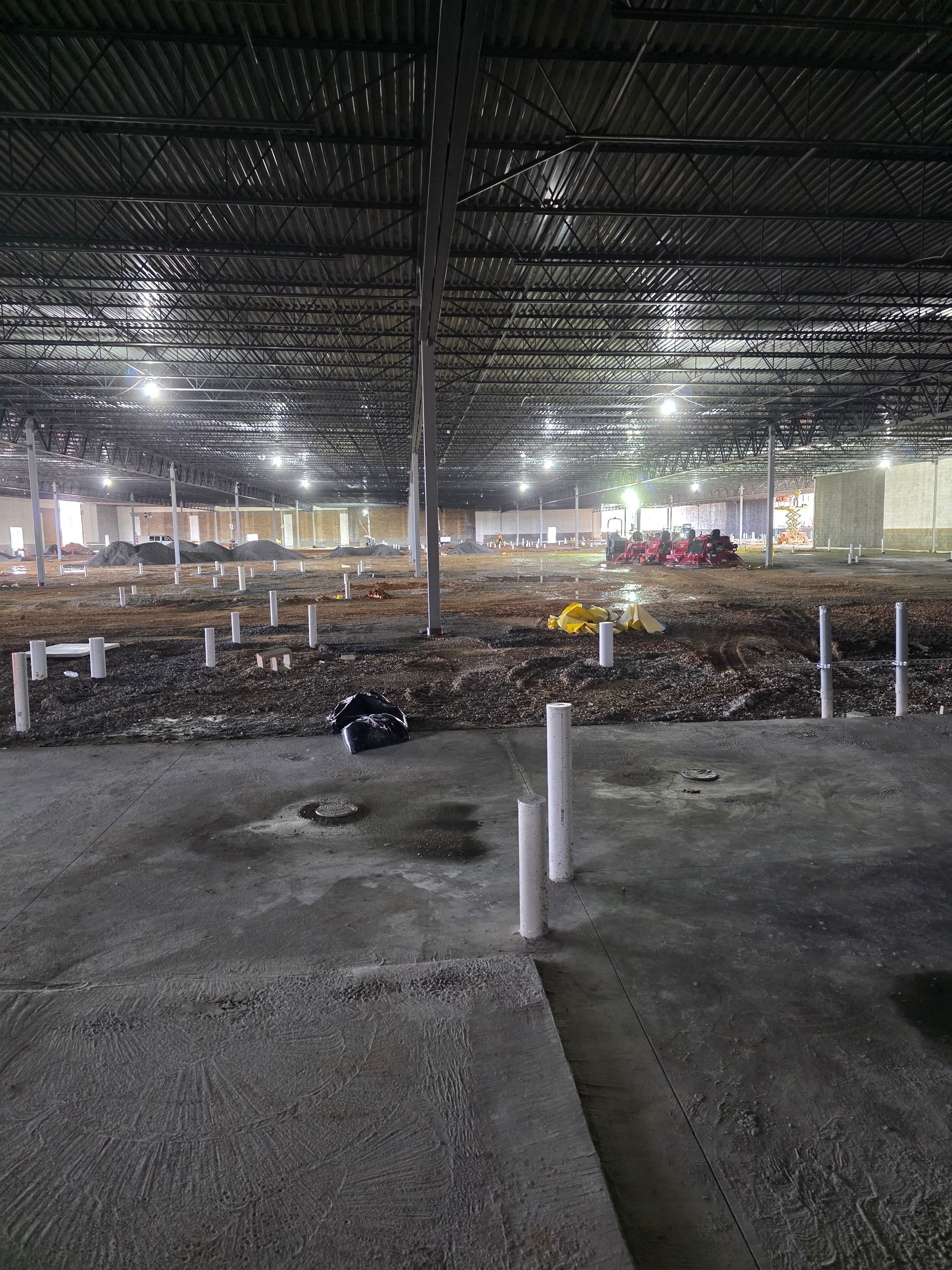 Interior of large unfinished building with exposed beams, pipes, and debris on the floor, lit by overhead lights.