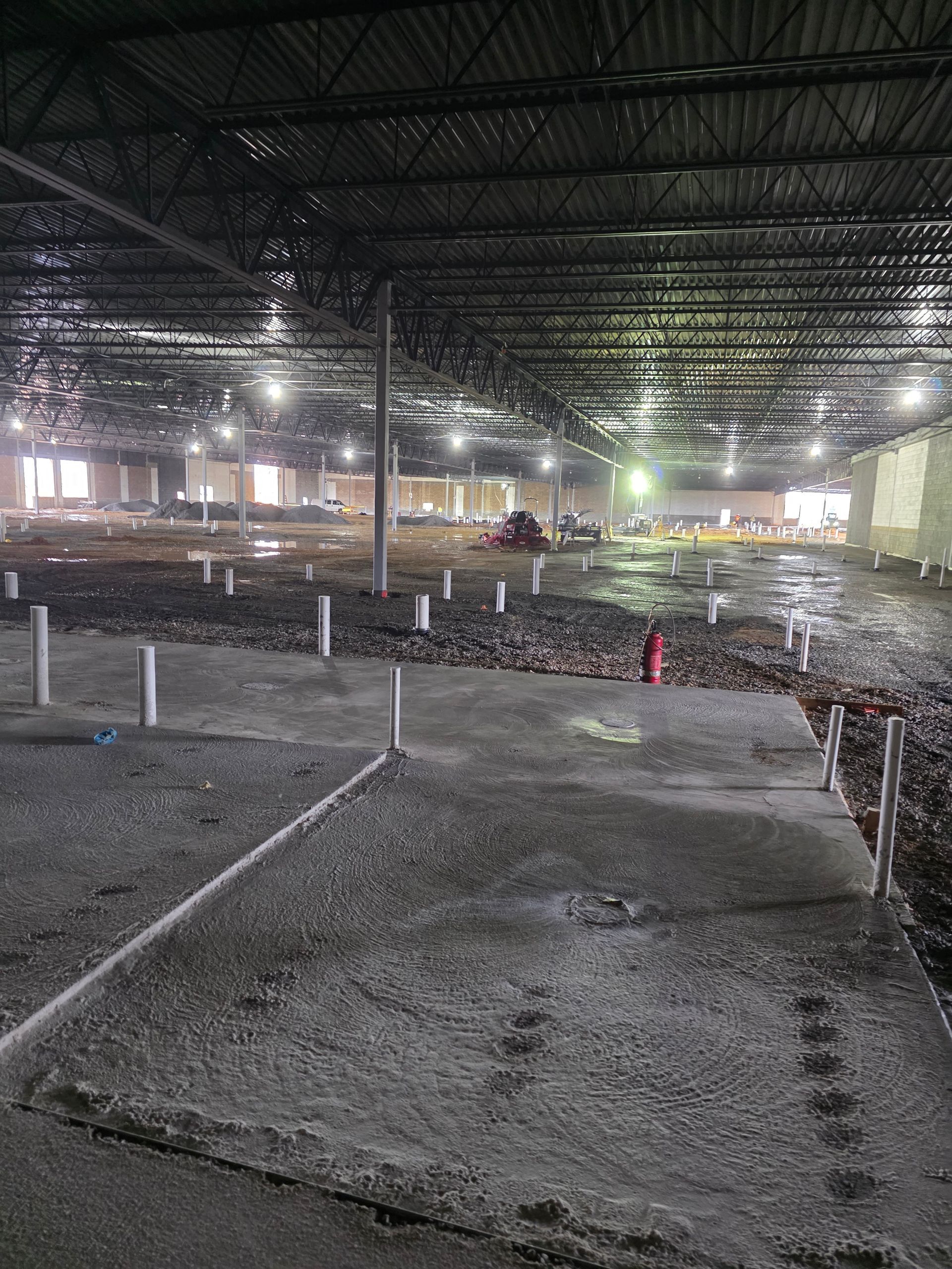 Interior of a large unfinished building with concrete flooring and plumbing pipes, under construction.