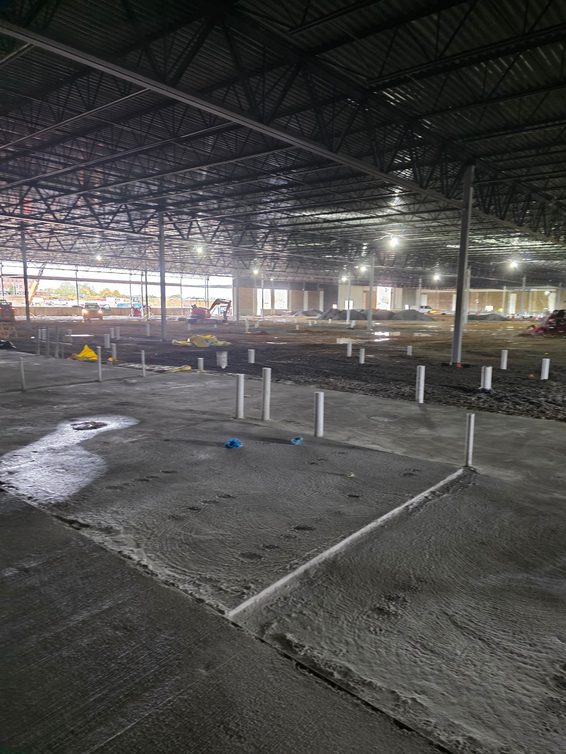 Interior of large building under construction with concrete floor, plumbing pipes, and metal roof.