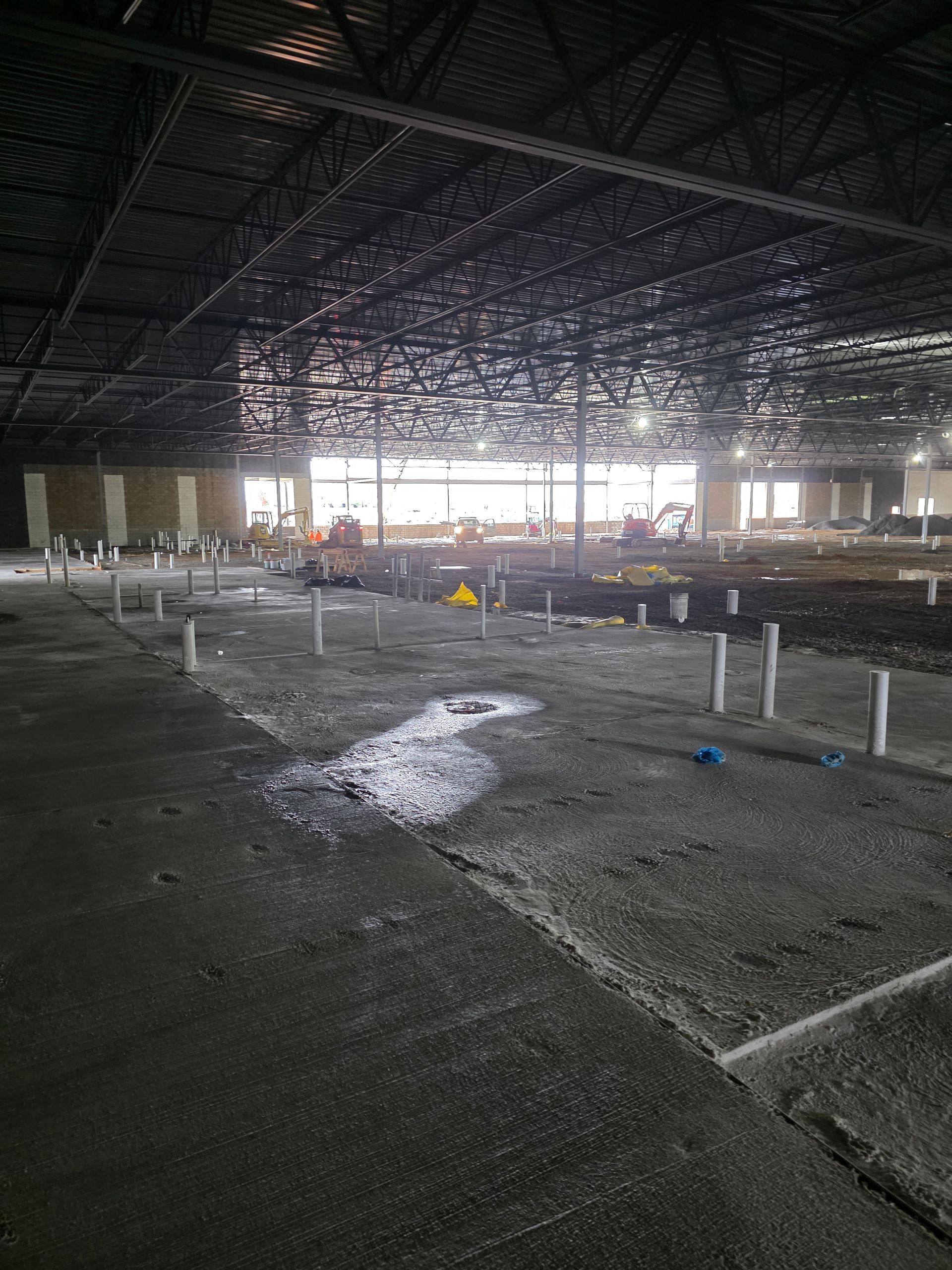 Interior of unfinished commercial building under construction; concrete floor, exposed ceiling, plumbing pipes, construction activity visible.