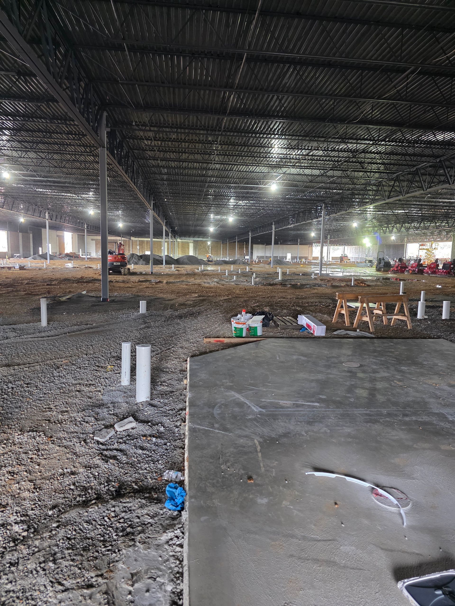 Interior view of a large warehouse under construction, with exposed beams, dirt floor, and a concrete section.