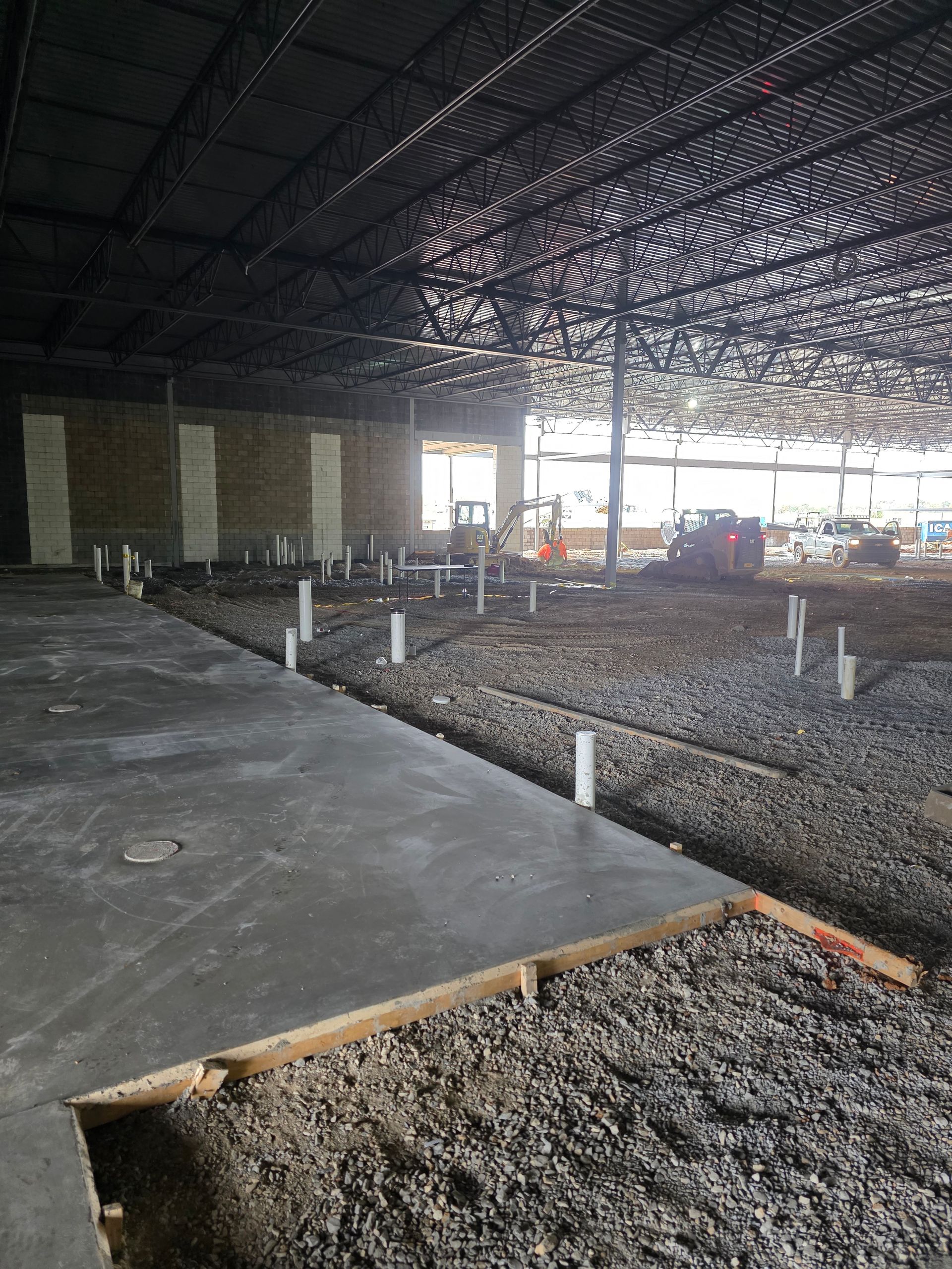 Interior of a building under construction. Concrete slab and gravel ground with plumbing pipes. Black metal frame overhead.