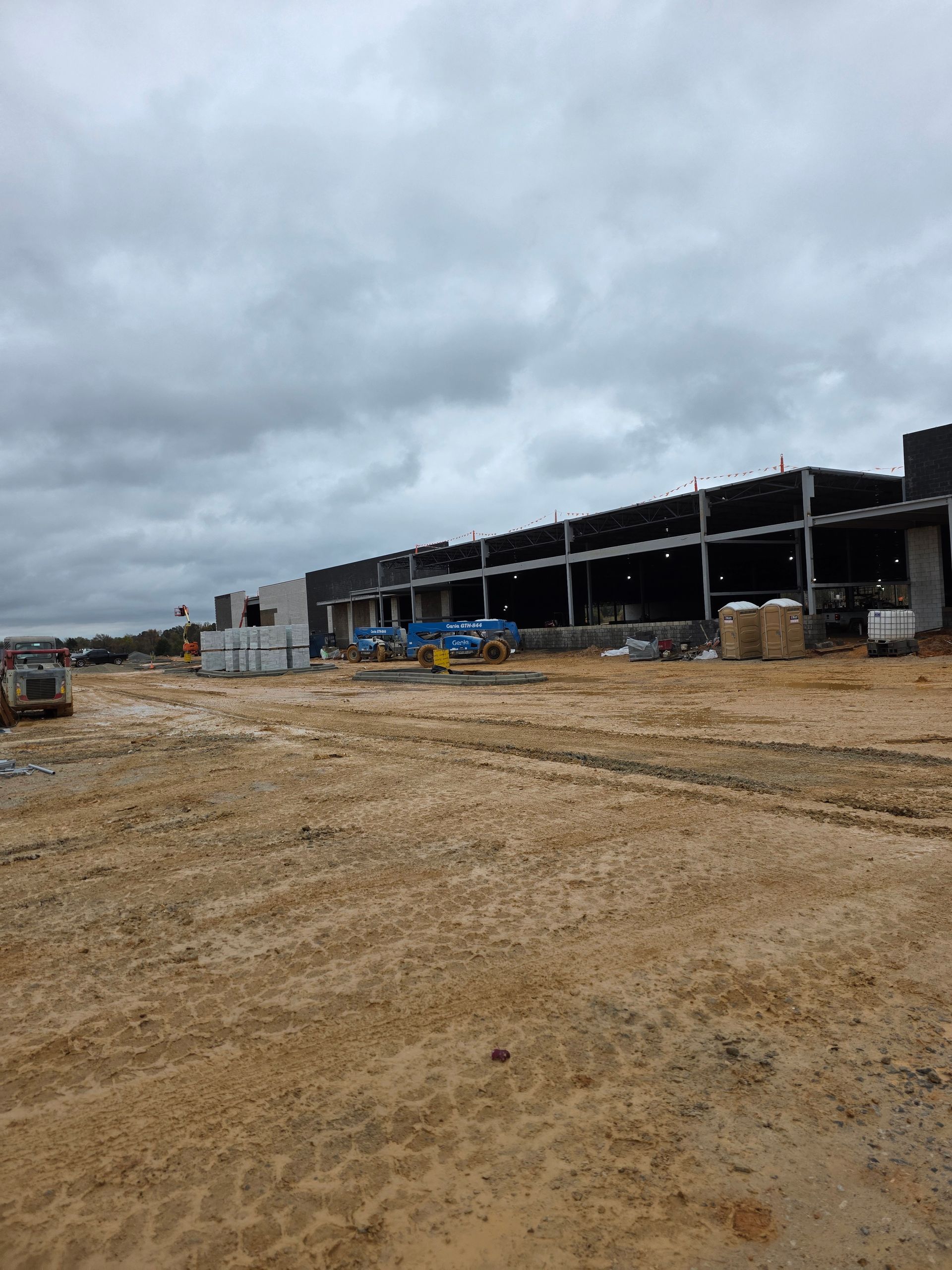 Construction site with large unfinished building under cloudy sky. Muddy ground with equipment.