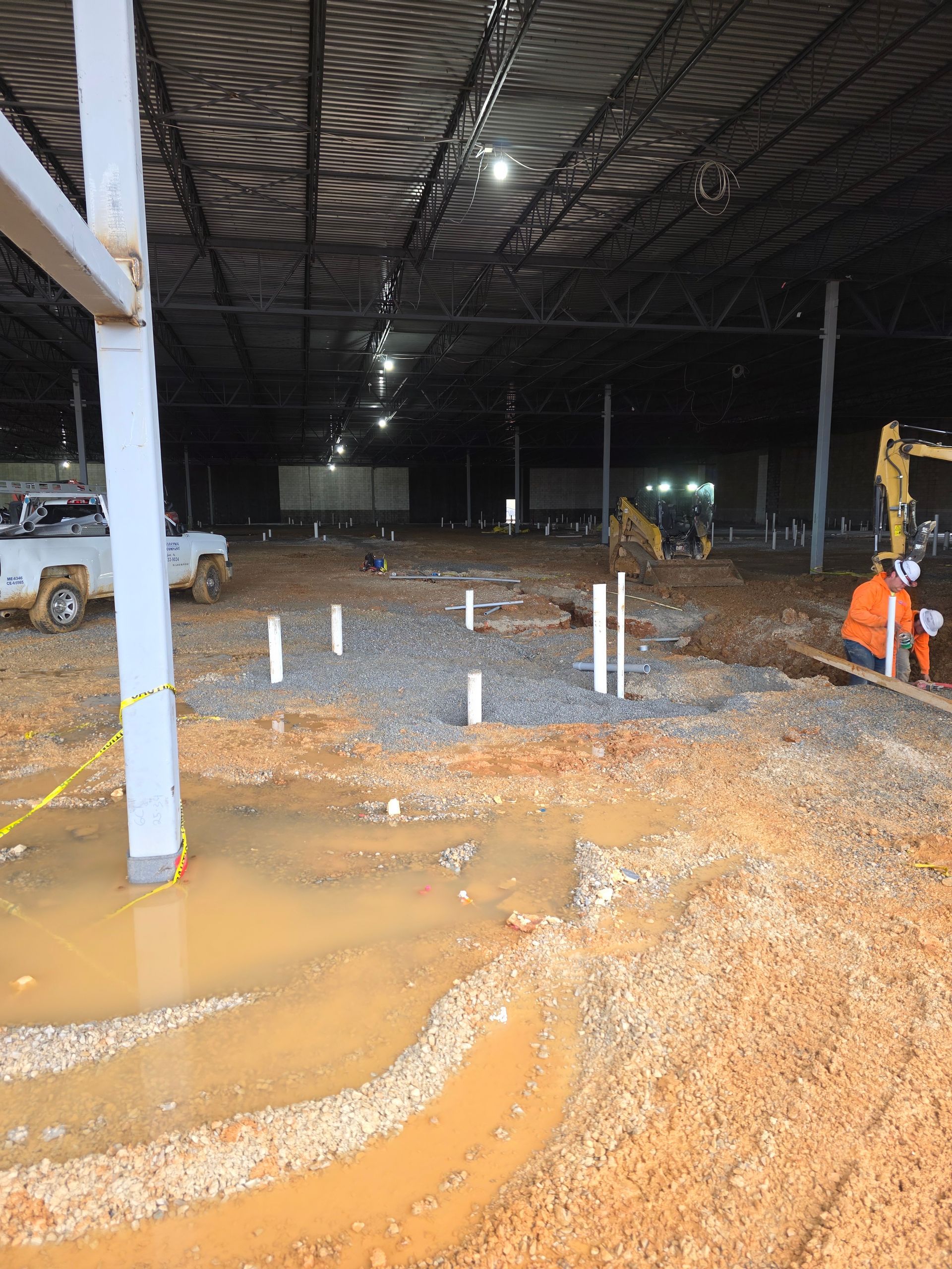 Construction site with standing water, gravel, pipes, a worker, and heavy machinery under a large dark structure.