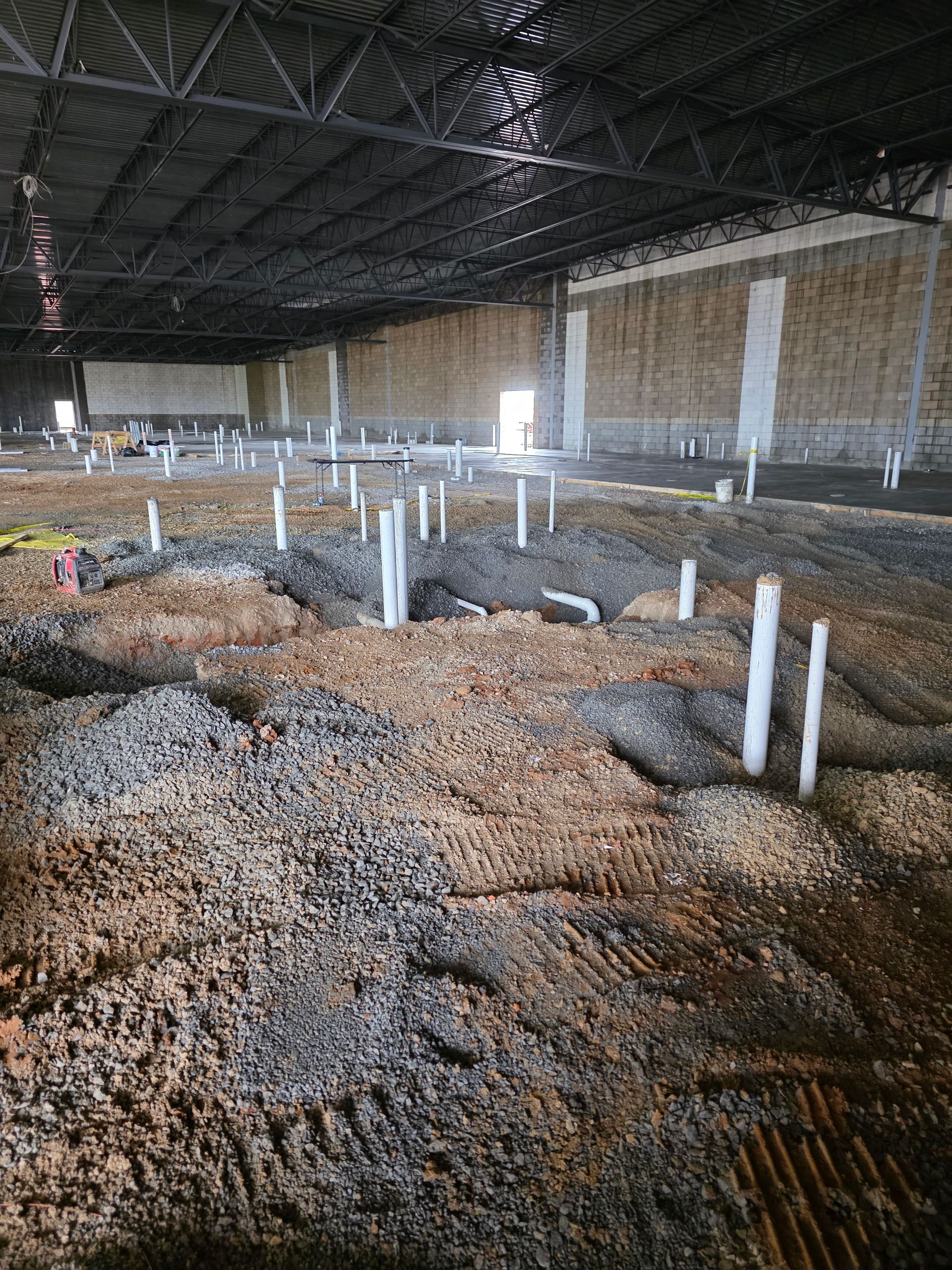 Interior of a large building under construction. Ground covered in dirt and pipes. Dark ceiling and walls.