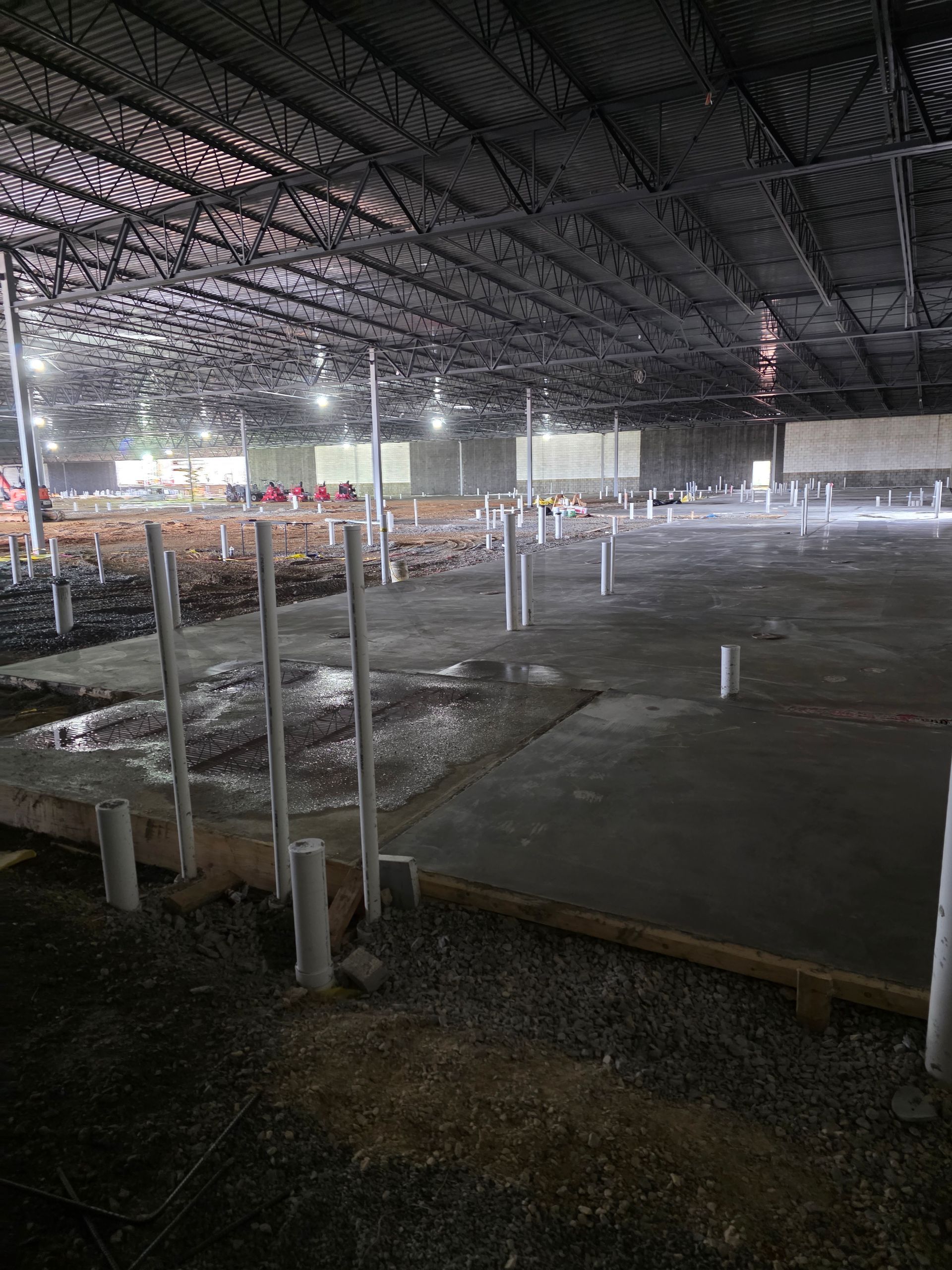 Interior of a large unfinished building with concrete flooring and plumbing pipes, under a metal ceiling with lights.