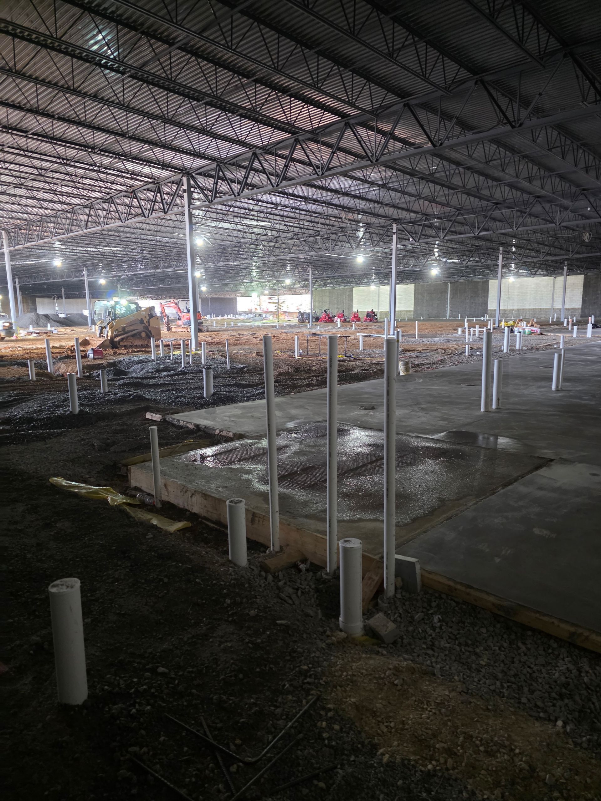 Interior of a large building under construction, showing plumbing pipes rising from a concrete floor.