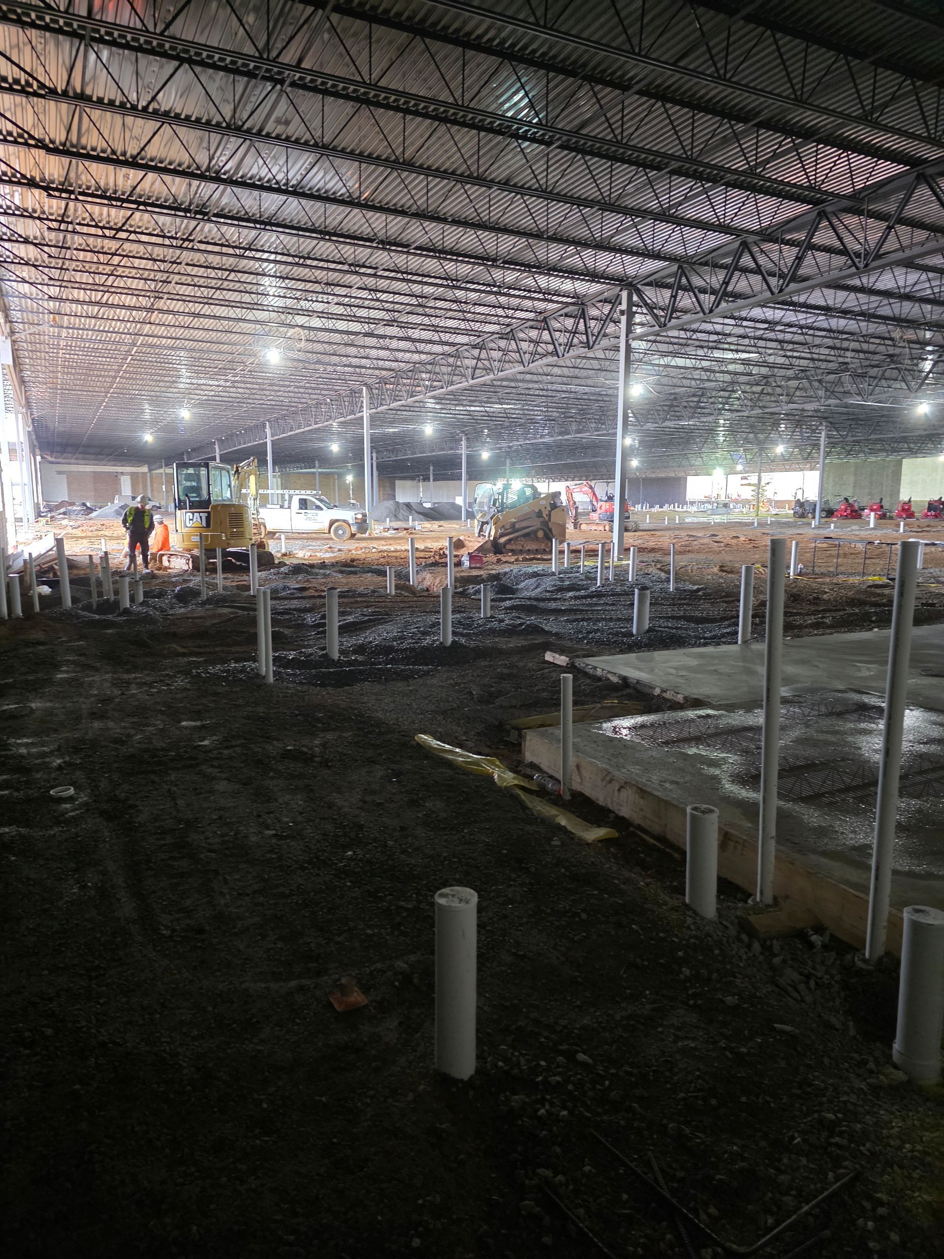 Interior construction site with exposed pipes, gravel, and heavy machinery under a metal ceiling.