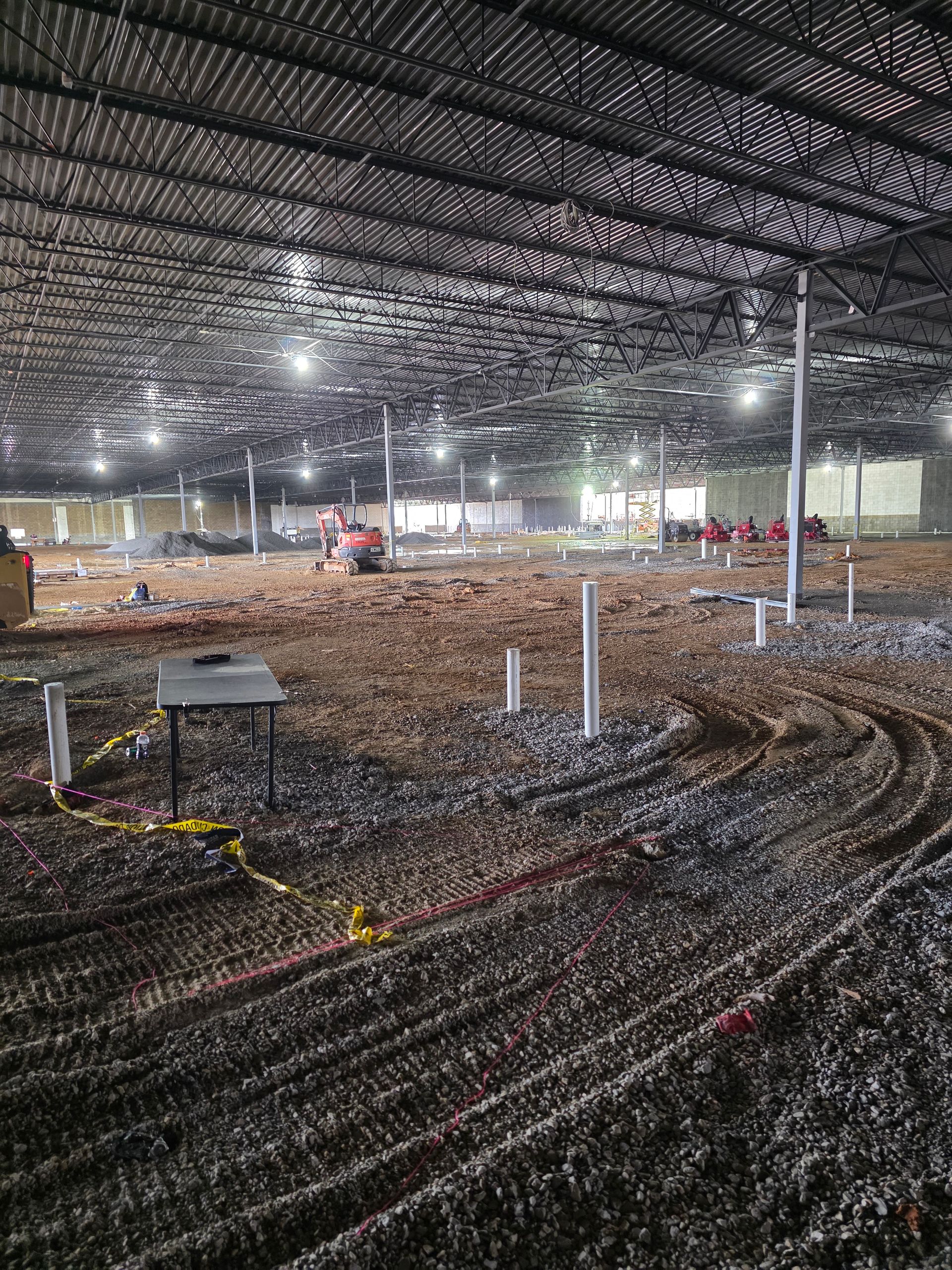 Construction site interior with exposed beams, dirt floor, plumbing pipes, and heavy machinery.