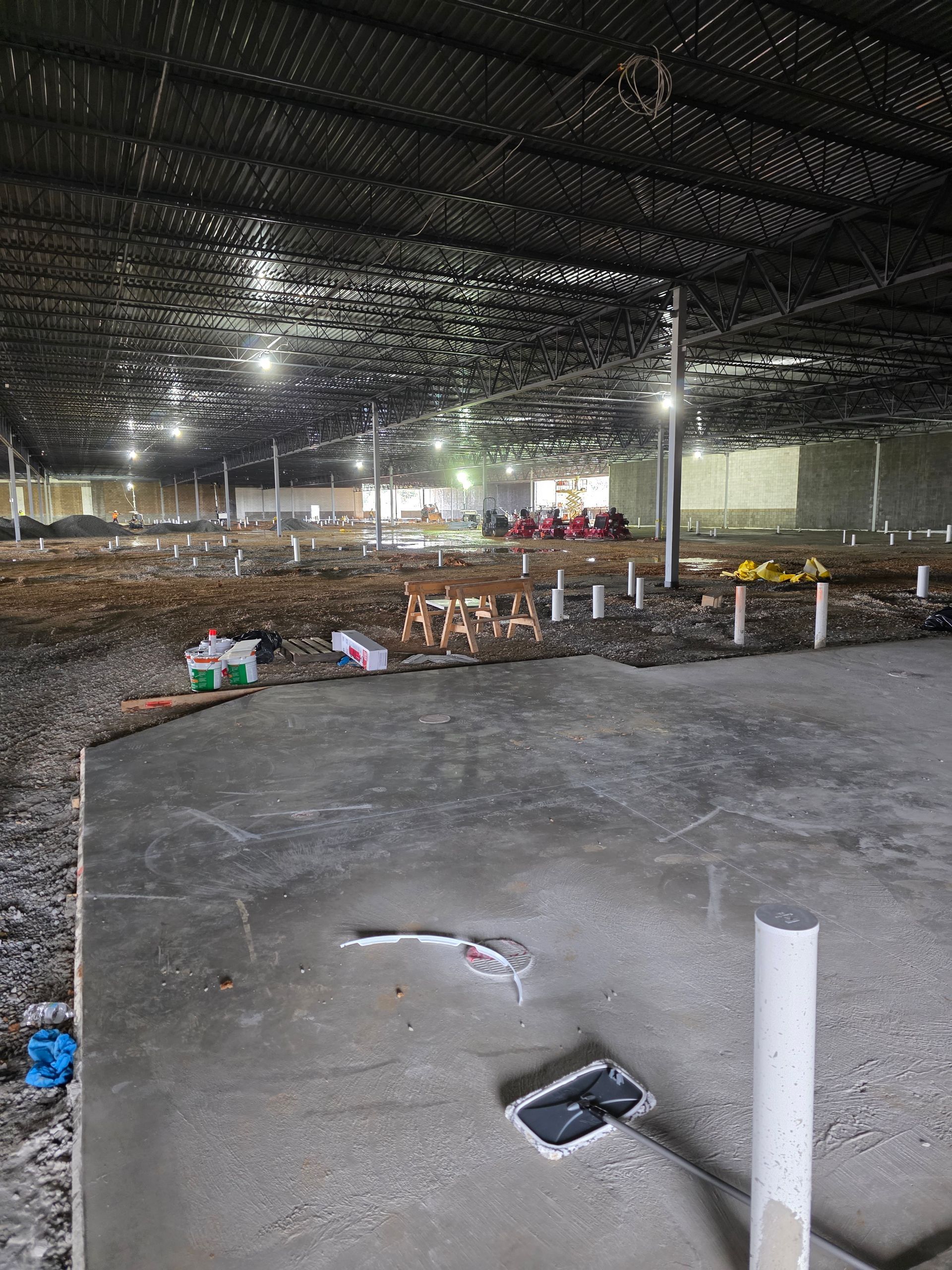 Interior of a large, unfinished building with concrete floor. Steel beams and overhead lighting.