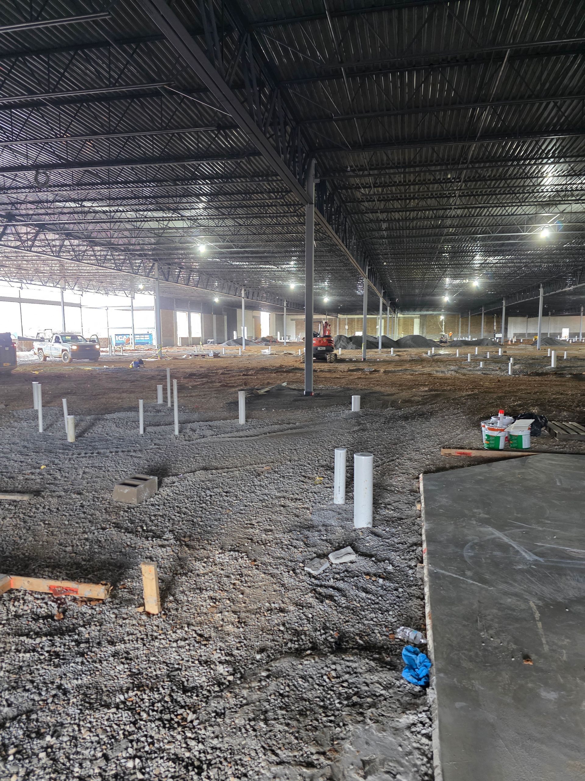 Interior of a large building under construction. Gravel ground with exposed plumbing pipes, steel beams, and ceiling lights.