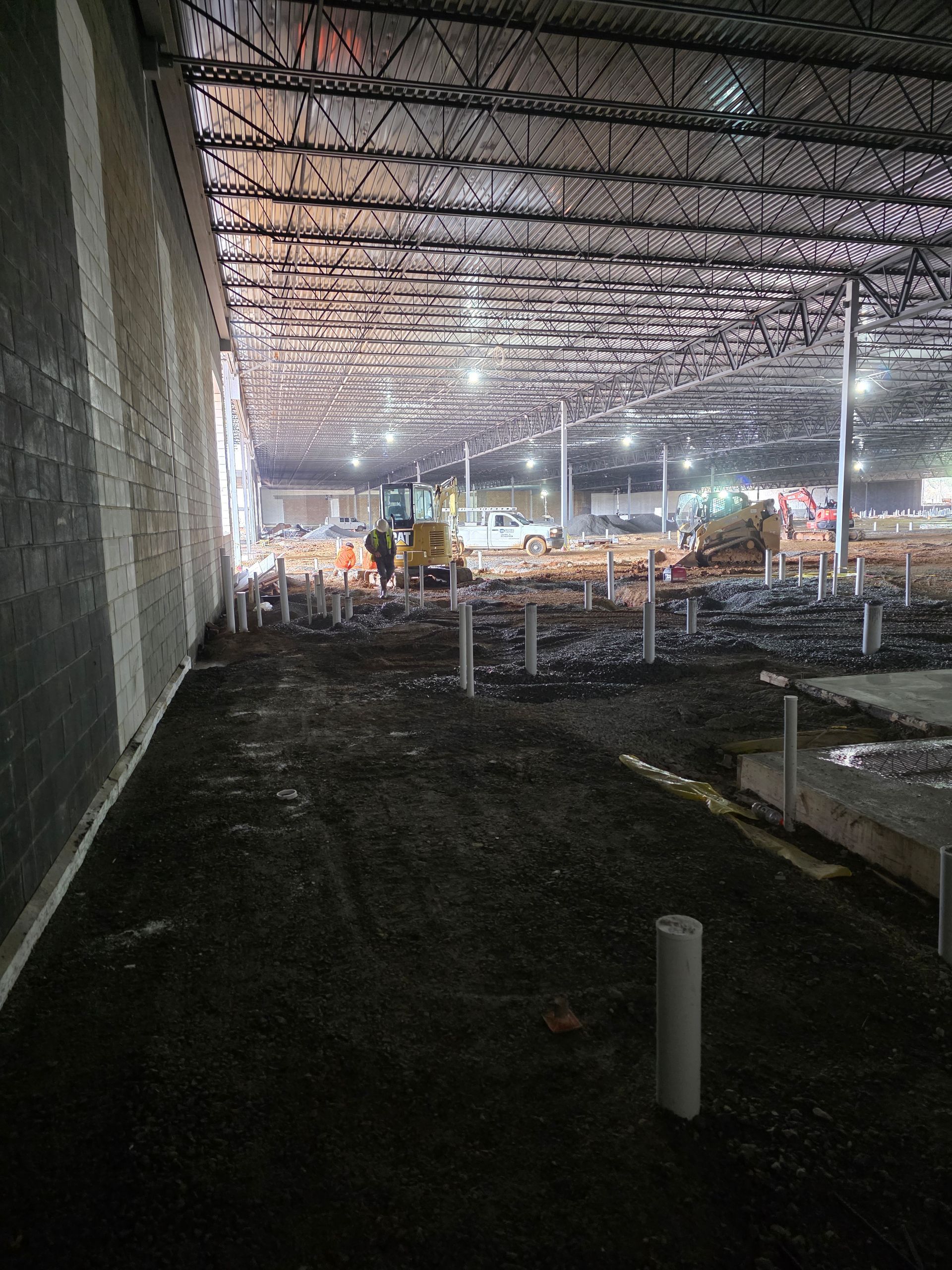 Interior of a building under construction, showing dirt floor, concrete columns, metal frame ceiling, and construction equipment.