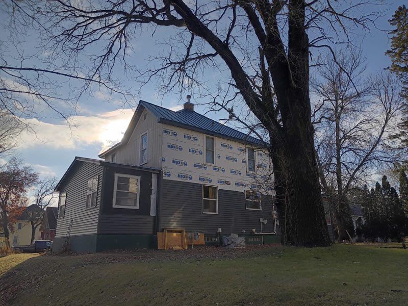 House under construction with blue roof, wrapped in white and blue protective covering, large tree on the right.