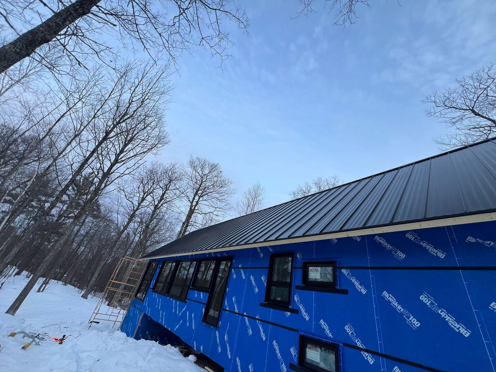 A partially constructed building with a black metal roof and blue exterior wrap in a snowy forest.
