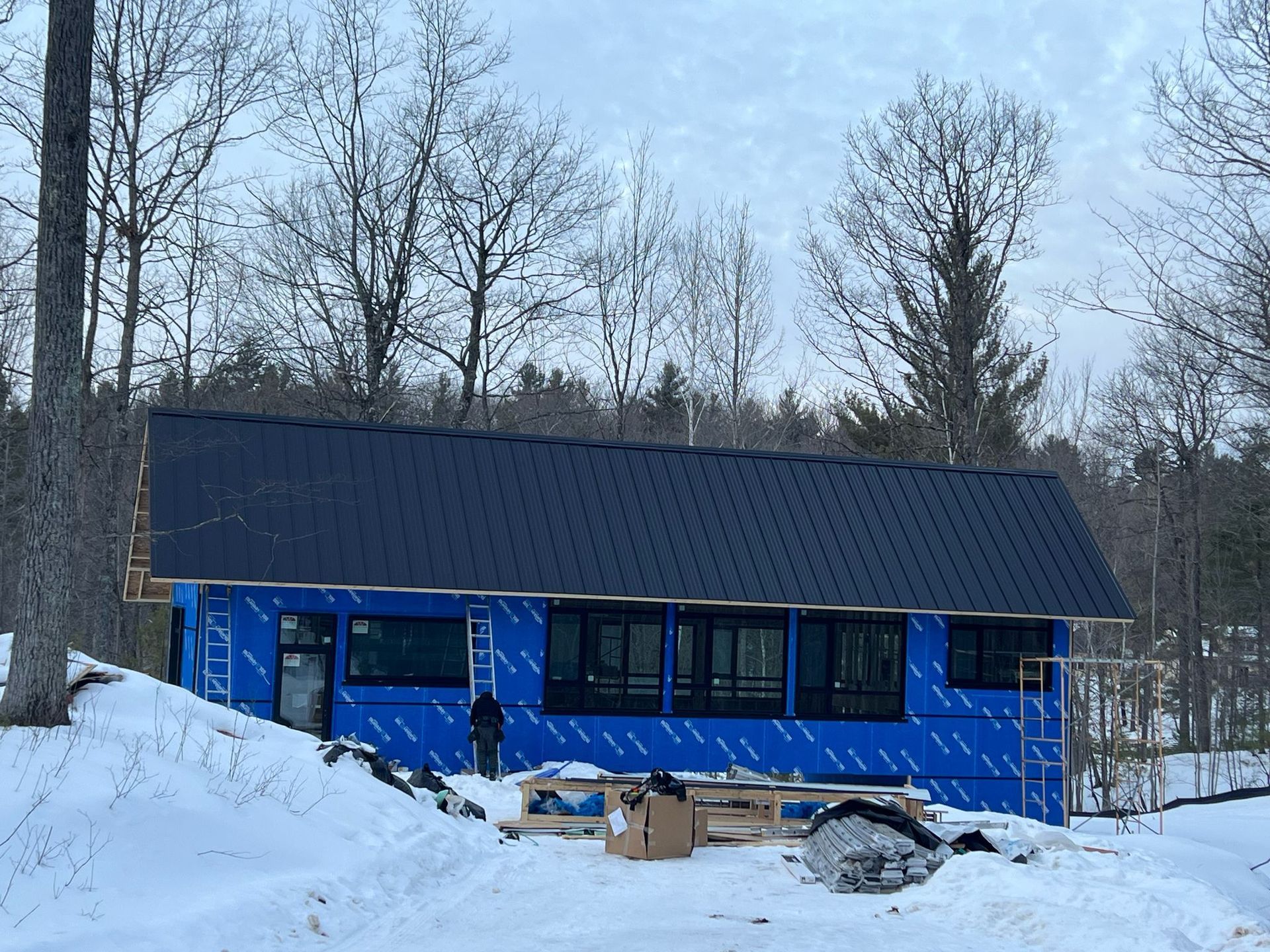 Cabin under construction with blue wrap, black roof, and snow.