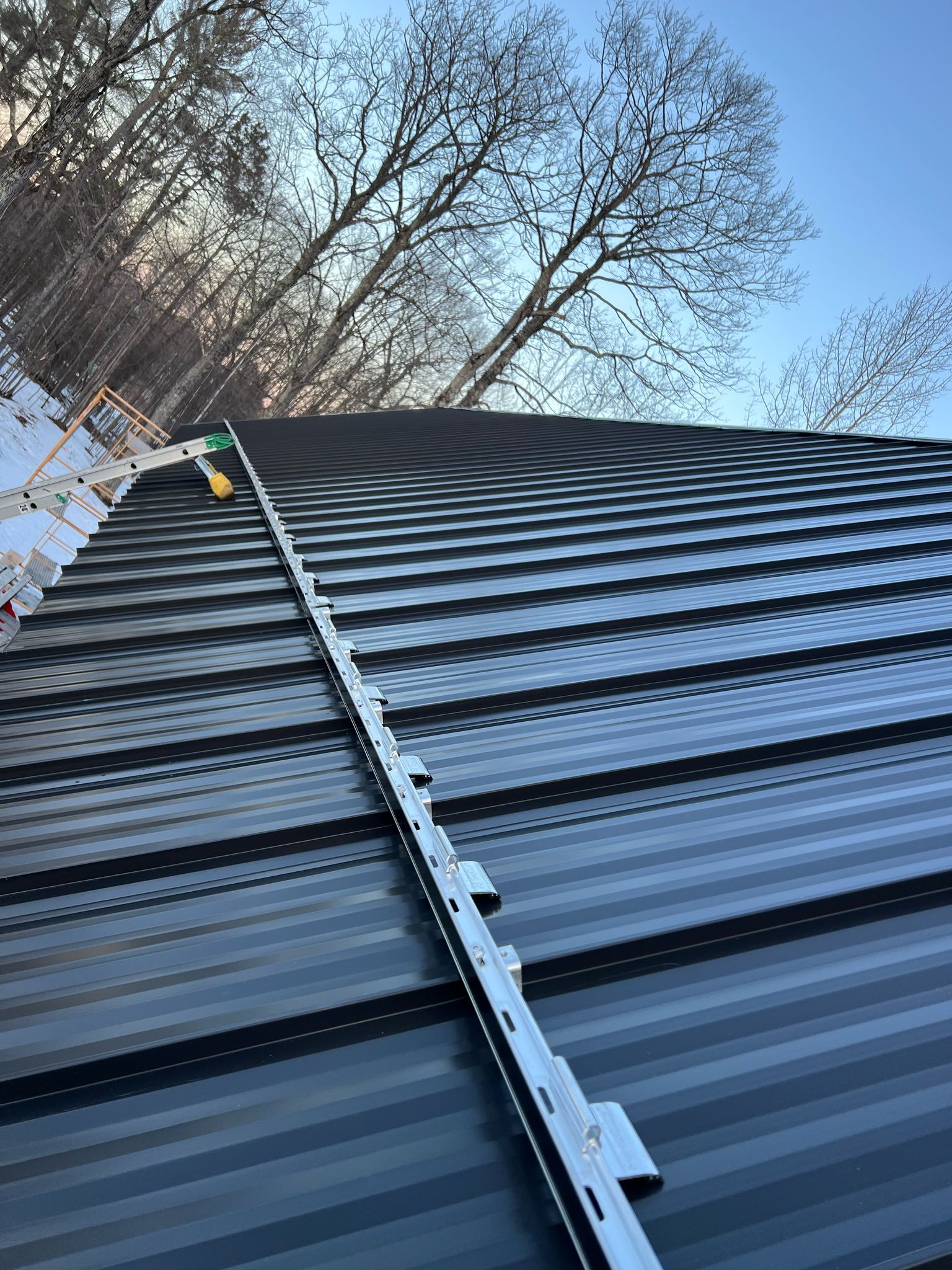 Black metal roof with safety railing and trees in the background under a blue sky.