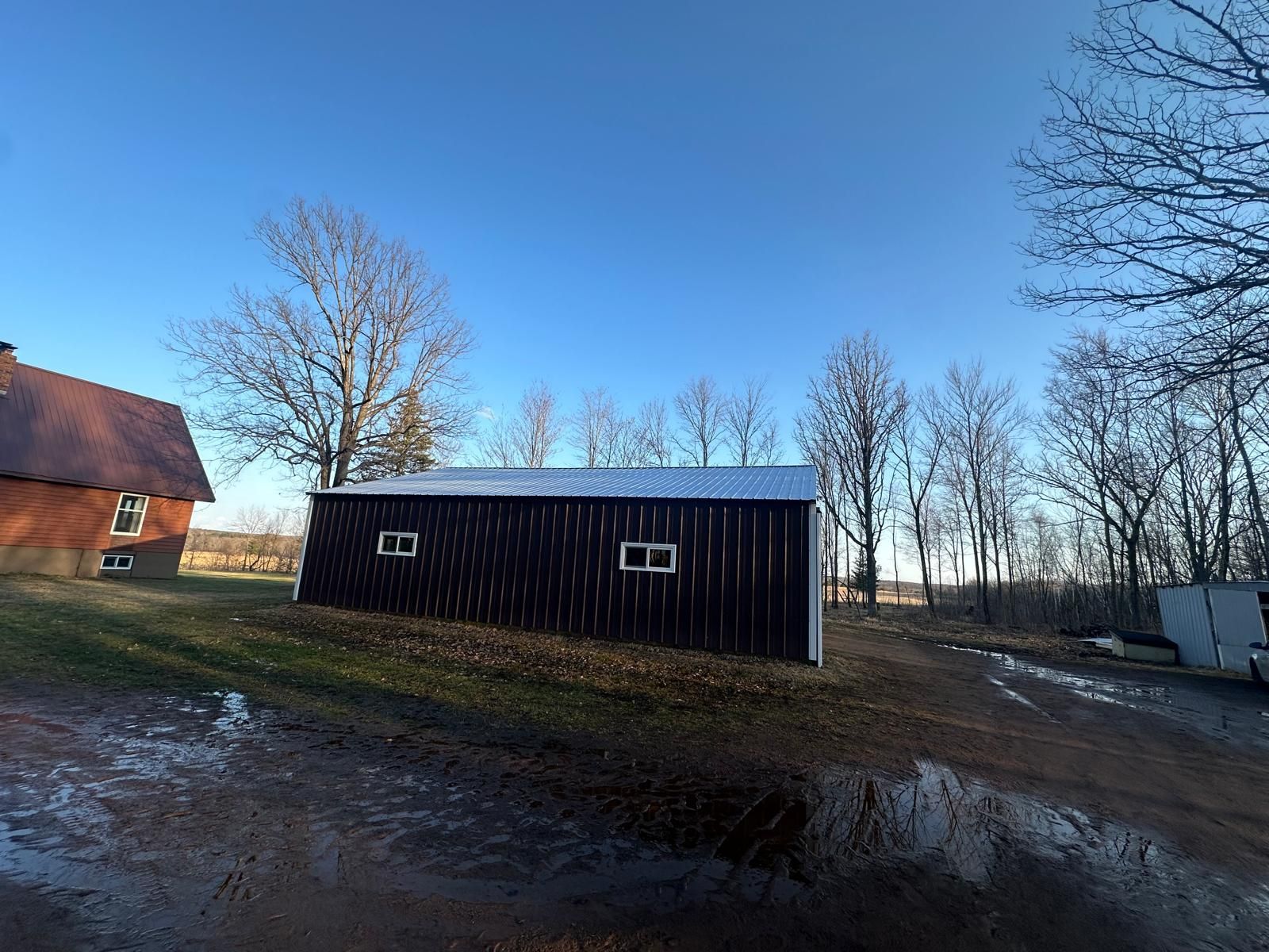 Long, dark-colored shed with white trim and two windows, set on a grassy hill with a blue sky.