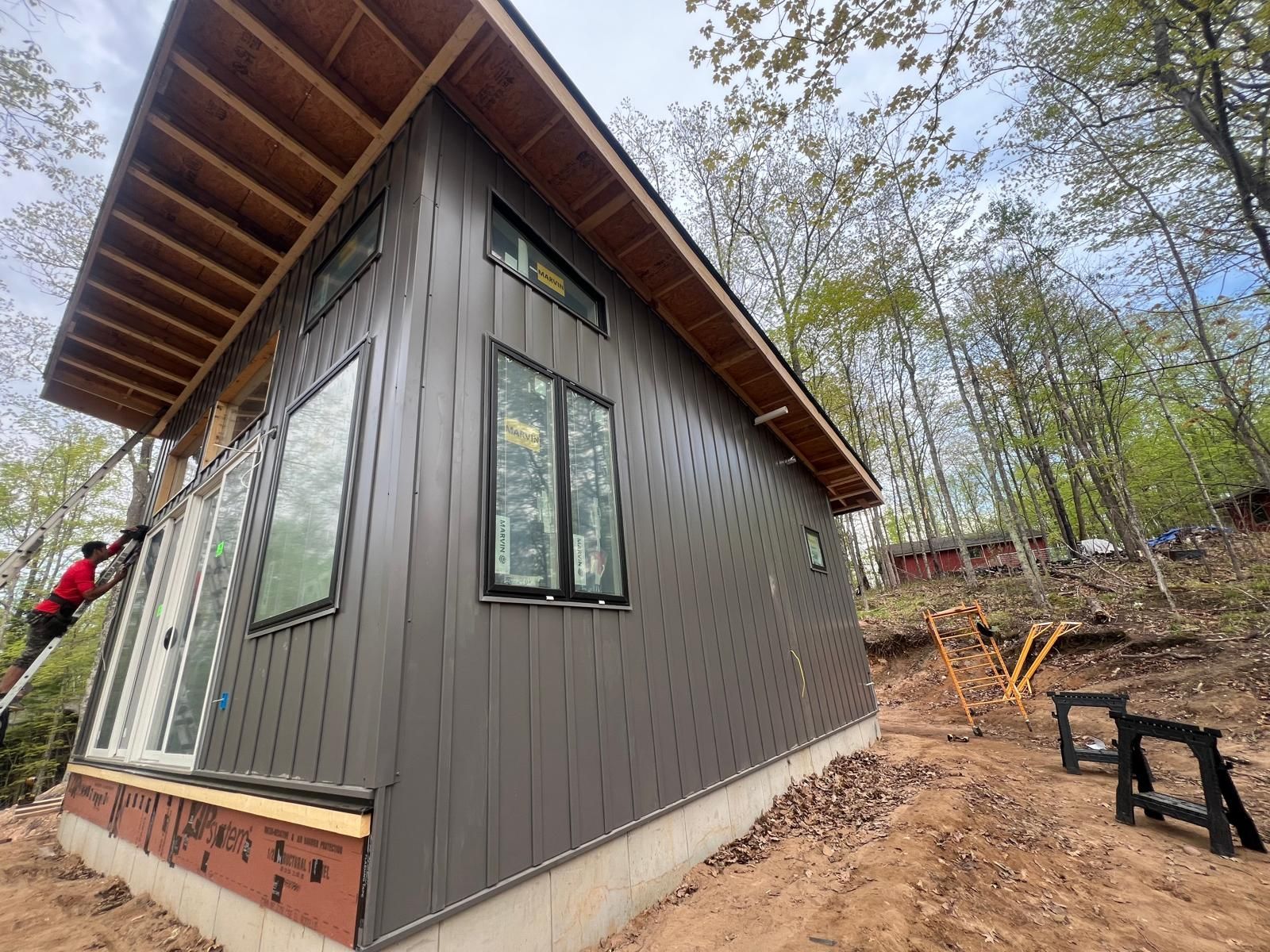 New cabin with dark gray siding, windows, and exposed wood roof trim; construction site.