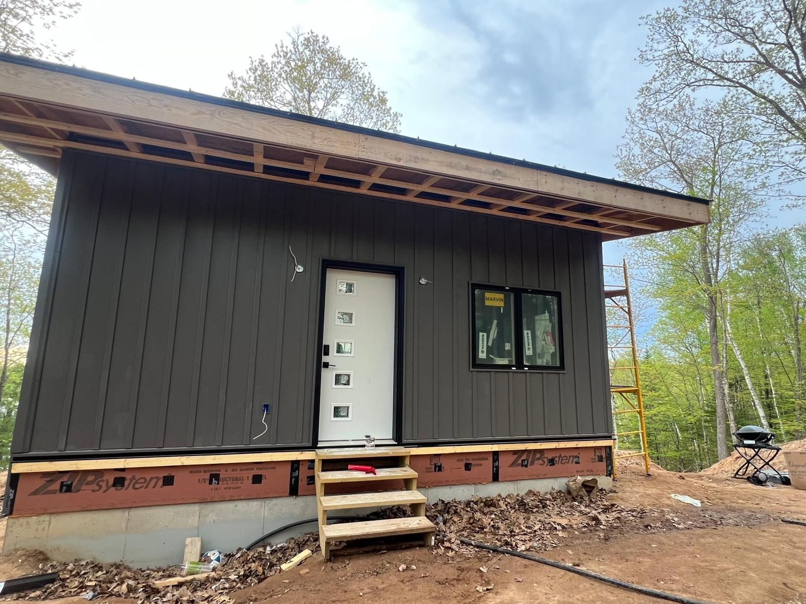 Brown-sided building under construction, white door, black window. Wooden stairs lead to the entrance. Scaffolding visible.