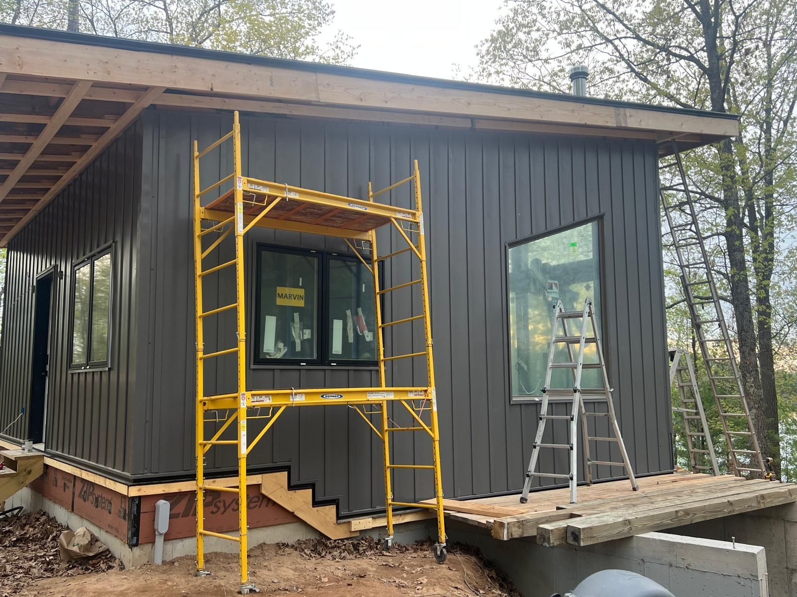 Building exterior under construction with dark gray siding, a yellow scaffold, and a small porch.