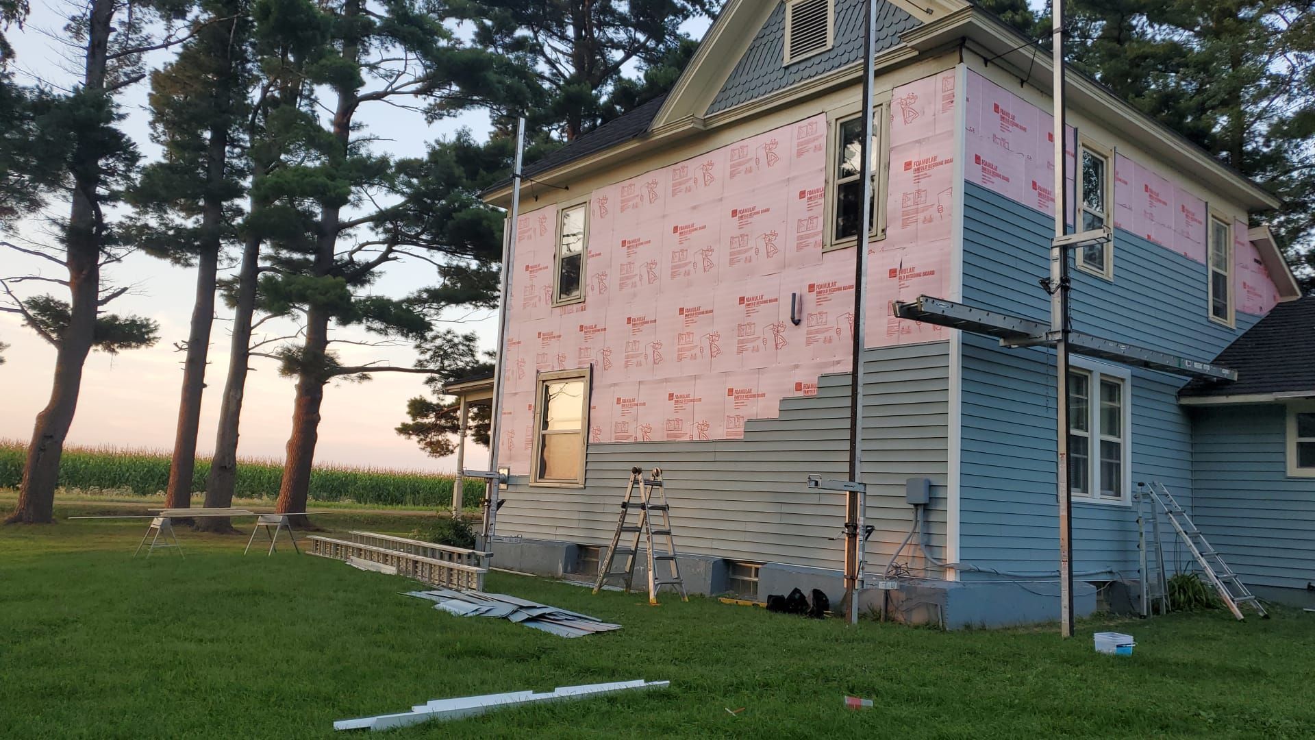 House exterior under renovation with scaffolding; pink insulation, blue siding, green lawn.