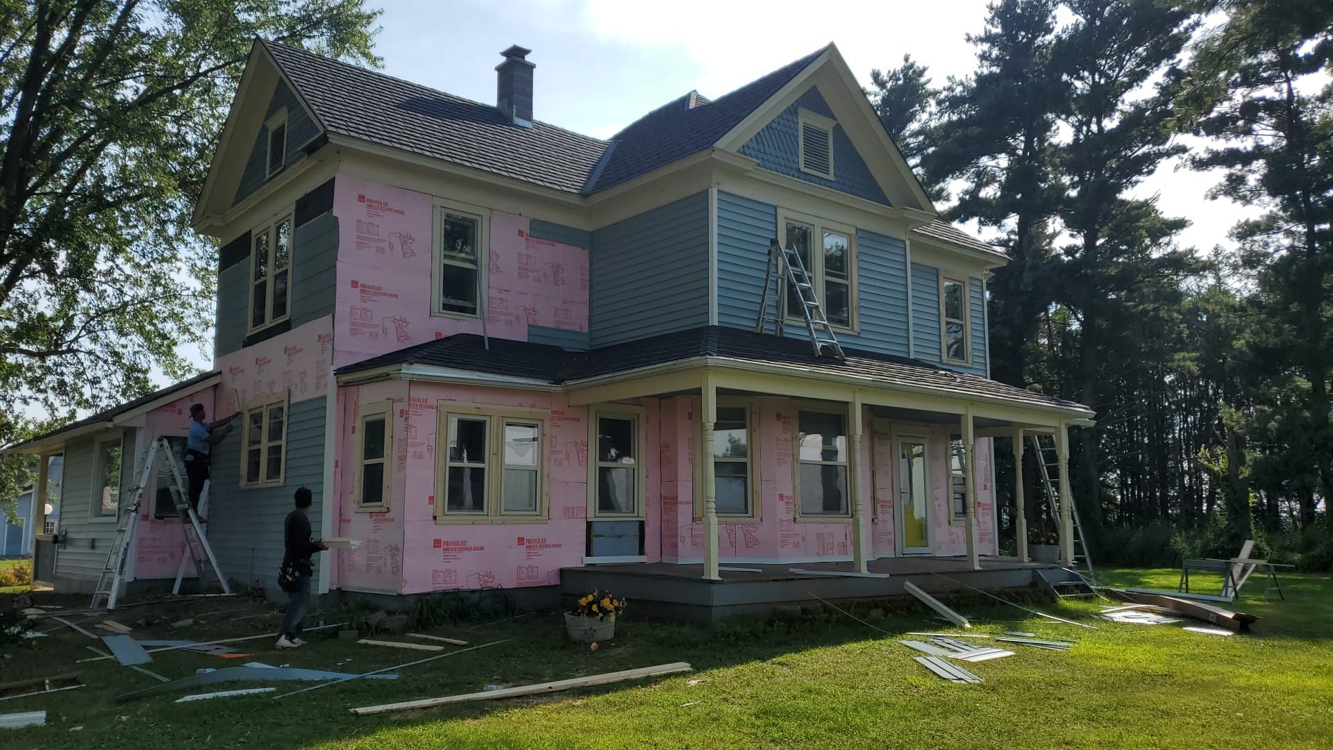 House under renovation with pink insulation and blue siding. Two workers on ladders.