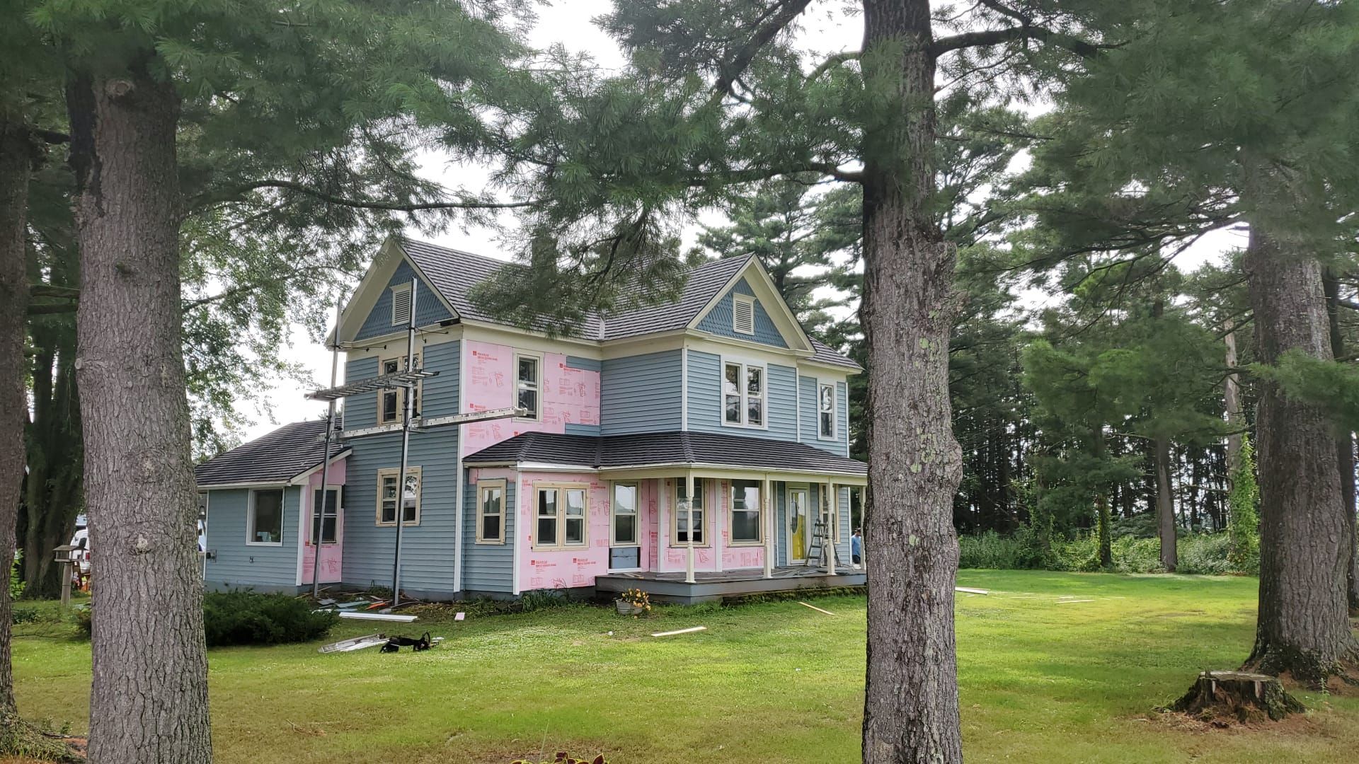 House under renovation, blue siding, pink insulation exposed, surrounded by trees and green grass.