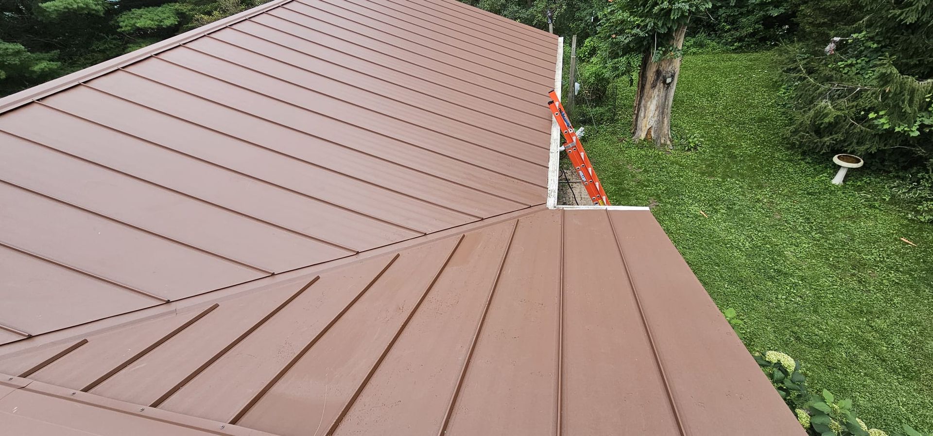 Brown metal roof of a house with visible lines, the edge of roof with a gutter and greenery background.