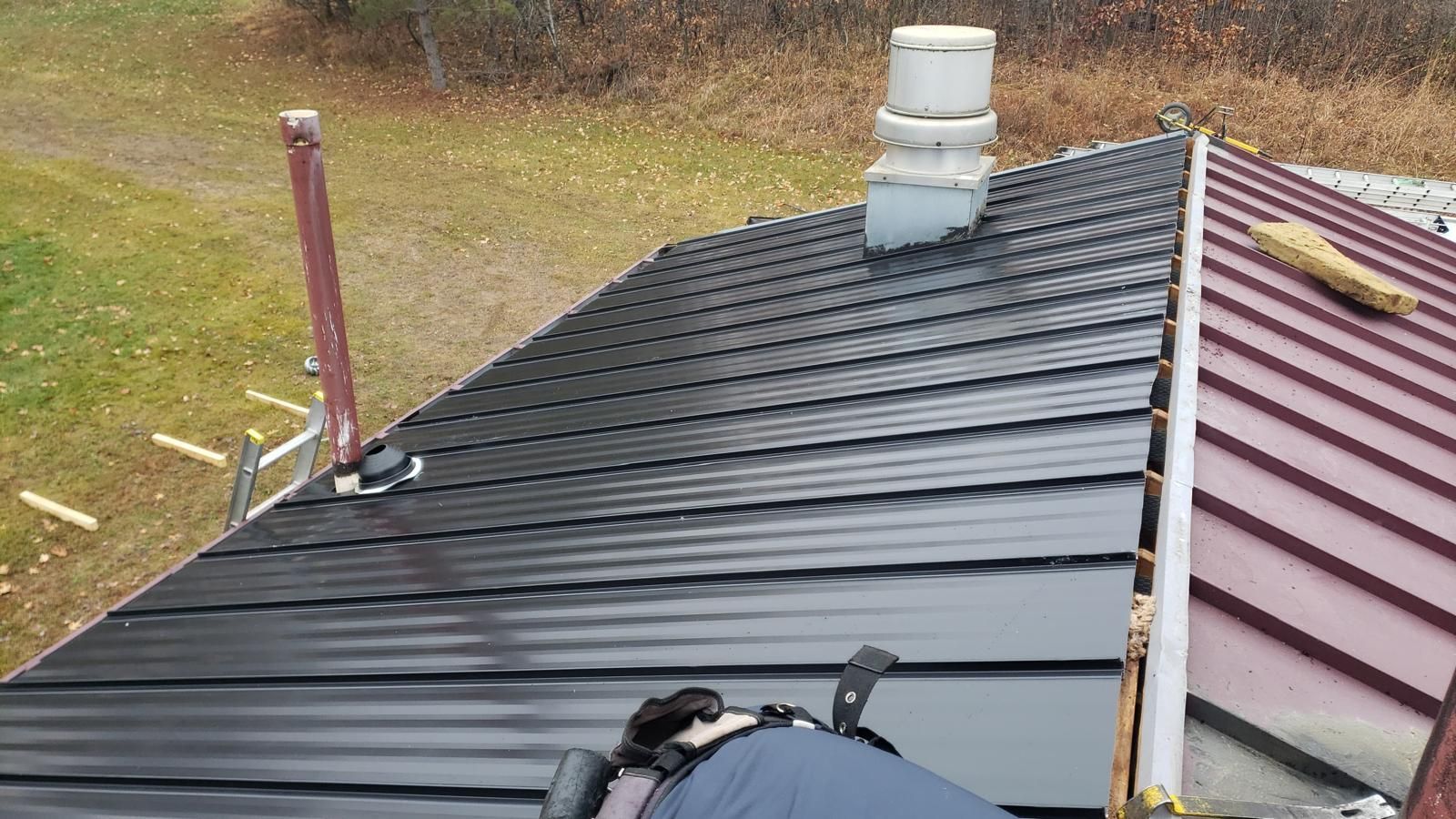 Person installing black metal roofing on a house; chimney and red roof section visible.