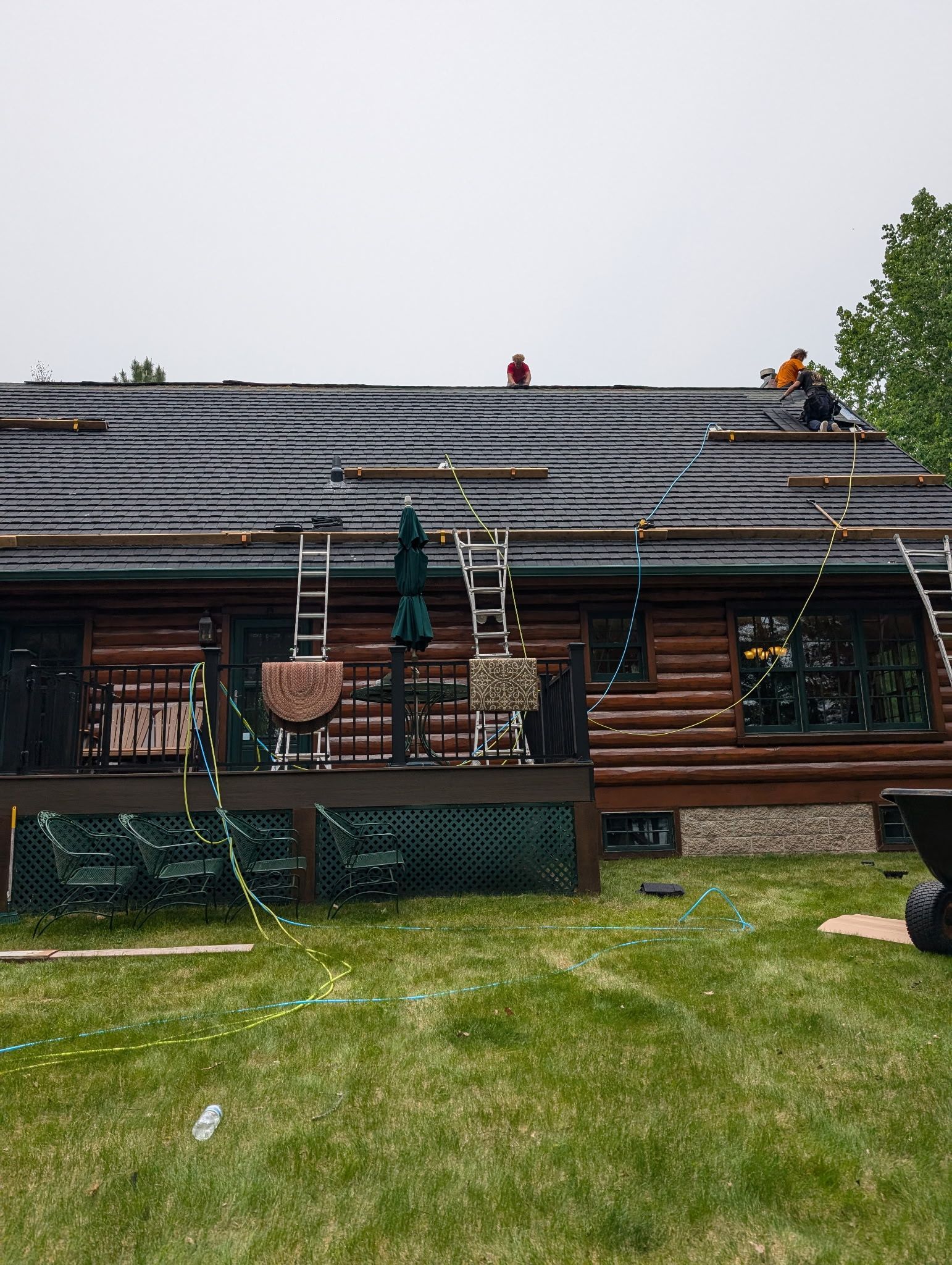 Workers replacing shingles on a log cabin roof, with ladders and a deck below.