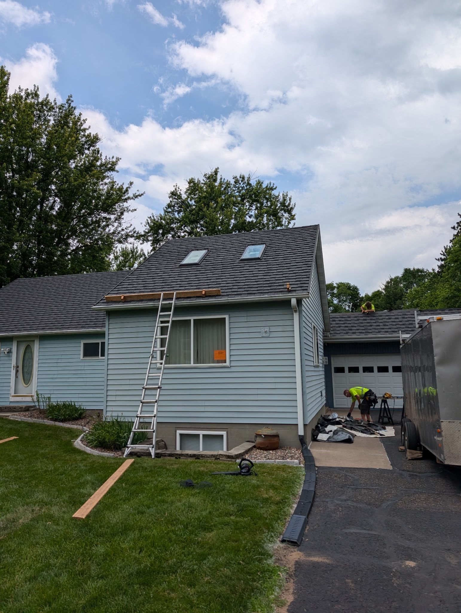 House with partially replaced dark roof; ladder, siding, trees, and workers near garage visible.