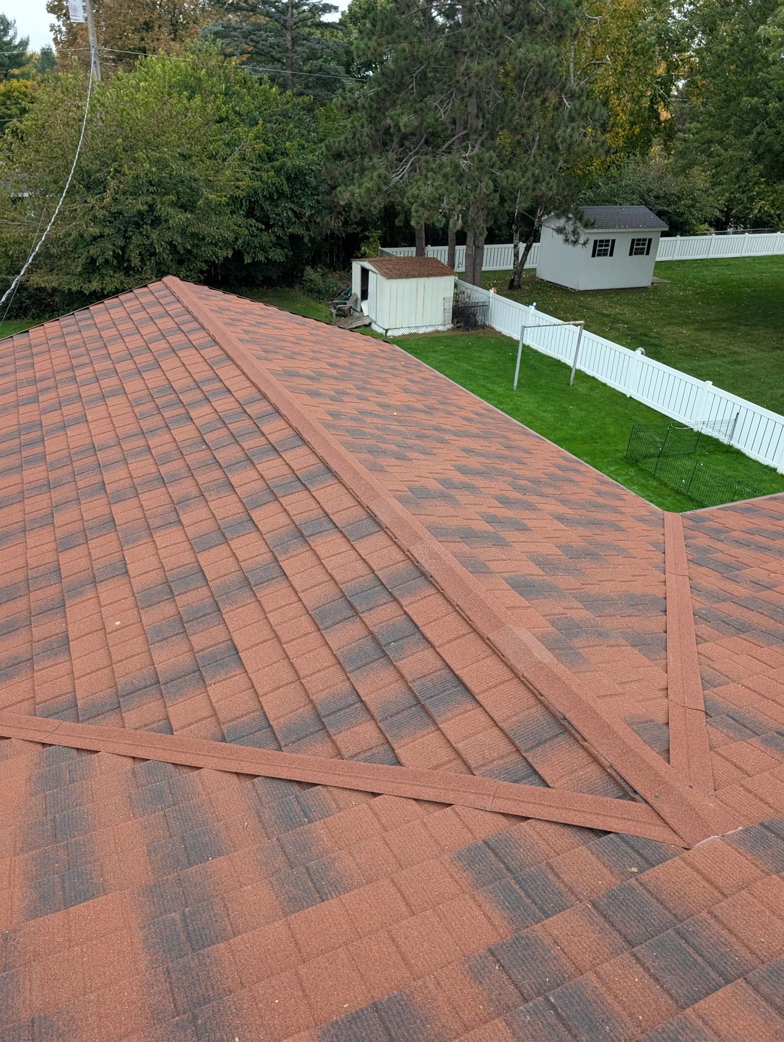 View of a newly shingled roof with a red and brown pattern. The roof overlooks a grassy yard and white fence.
