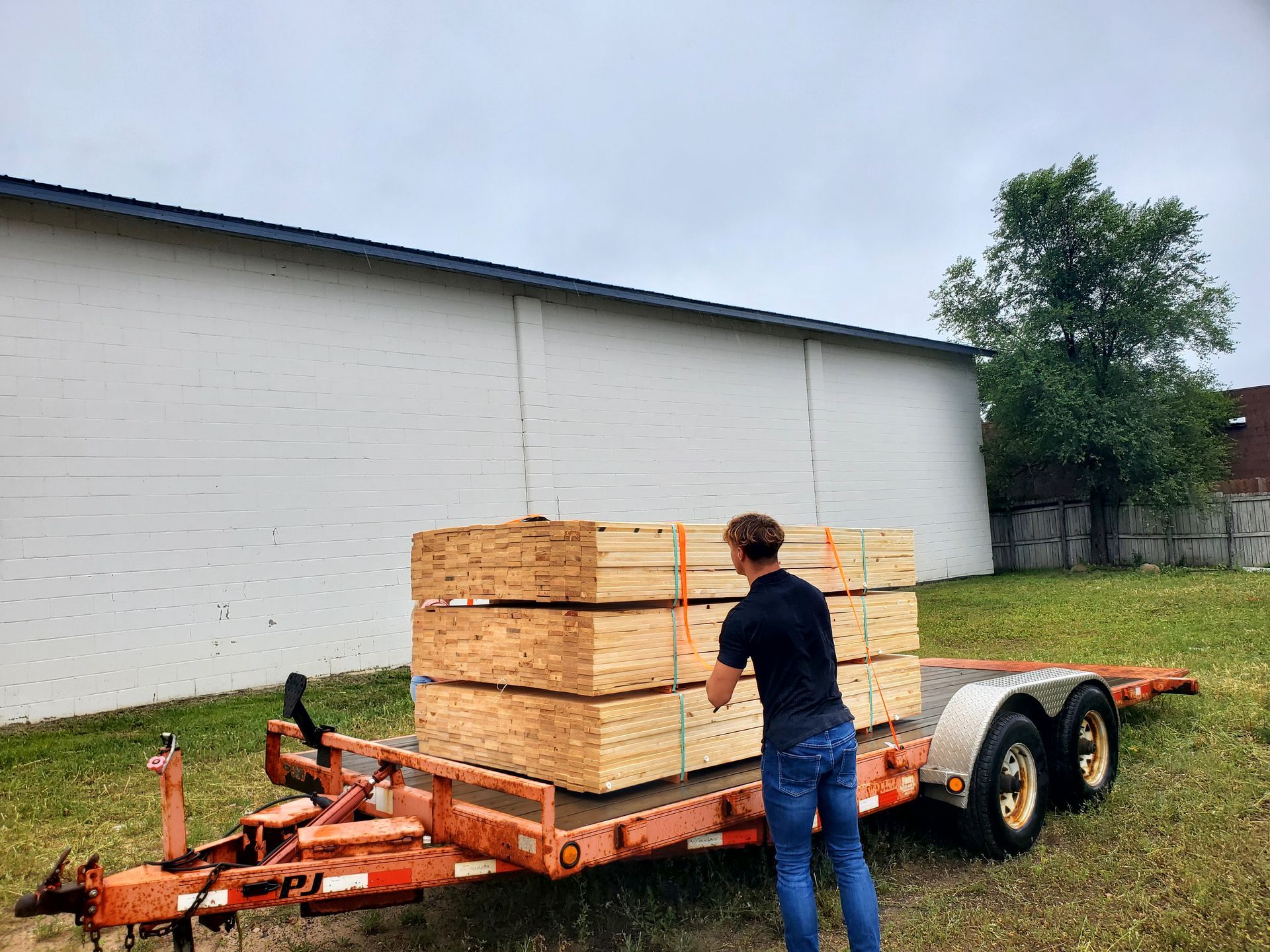 Person securing lumber on an orange trailer in front of a white building on a cloudy day.