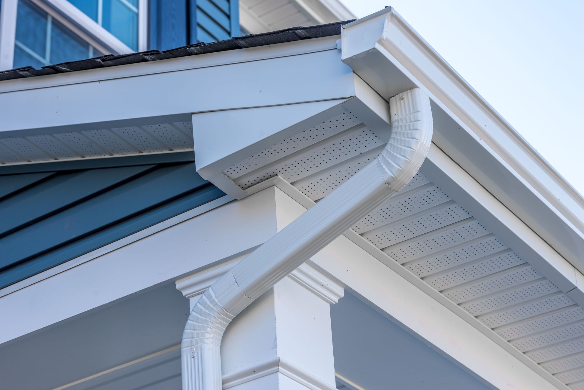 White gutters and downspout on a light blue house, against a clear sky.