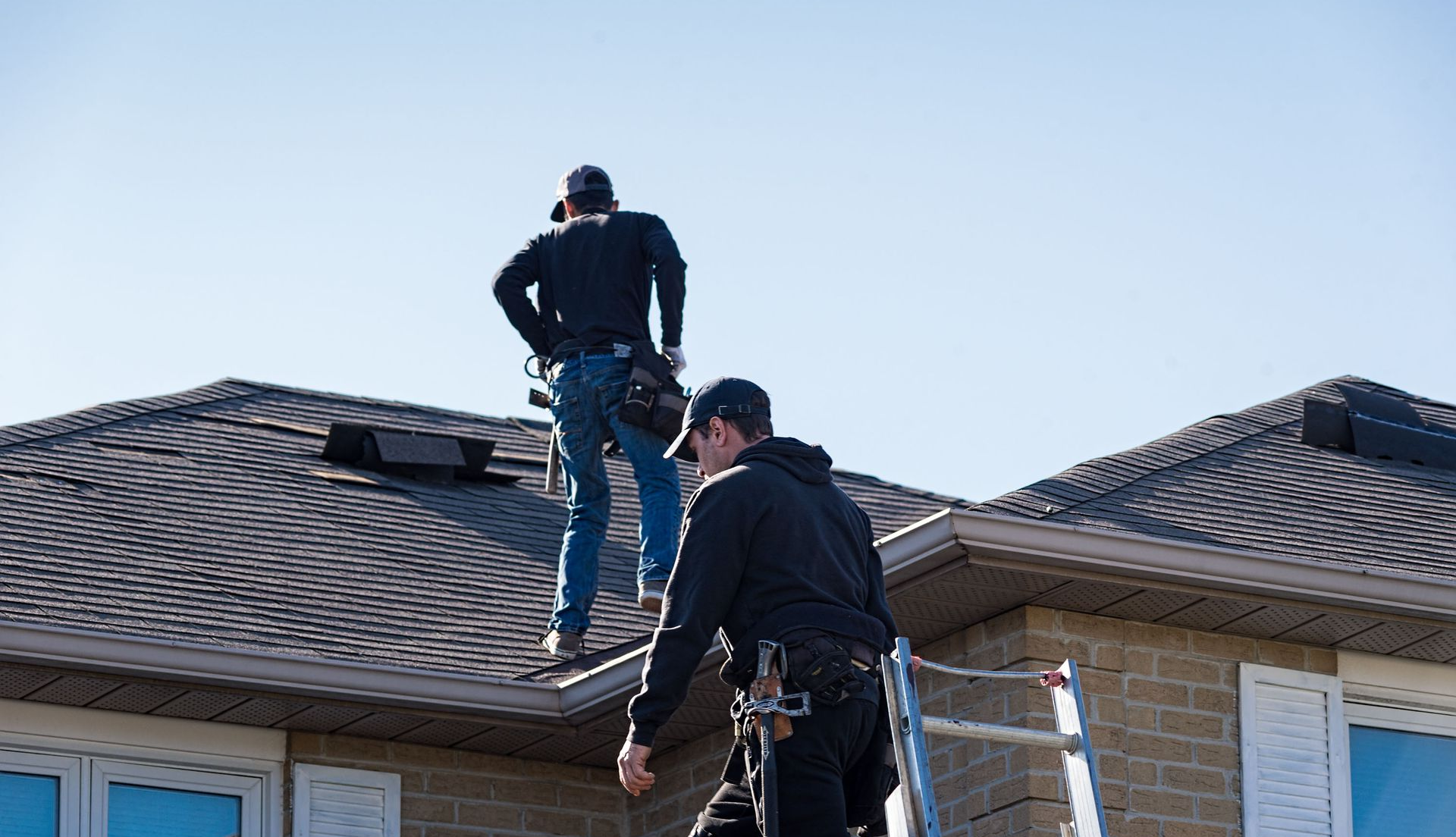 Two workers on a dark shingle roof, one on the ground, one standing, wearing safety harnesses and tool belts.