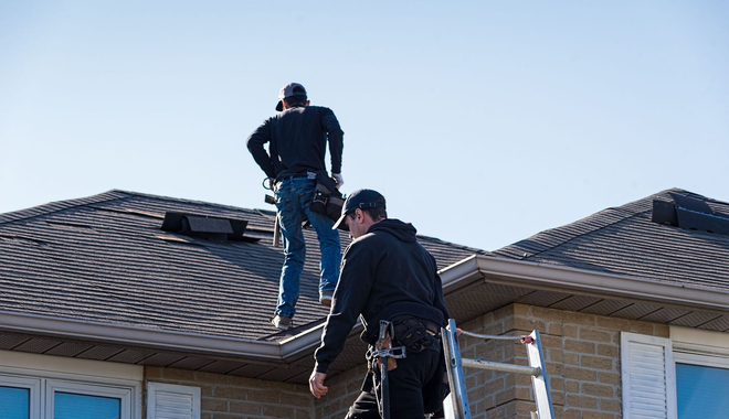 Two workers on a dark shingle roof, one on the ground, one standing, wearing safety harnesses and tool belts.