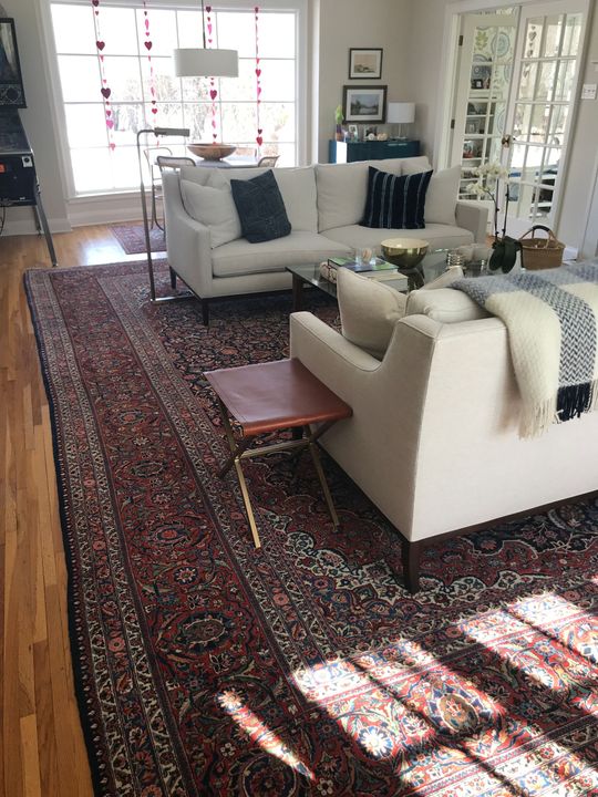 Living room with white sofa and chairs on a red Persian rug, sunlight through window.