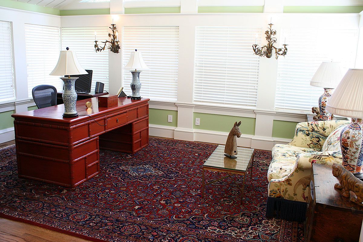 A formal office with a red desk, floral couch, area rug, and window blinds
