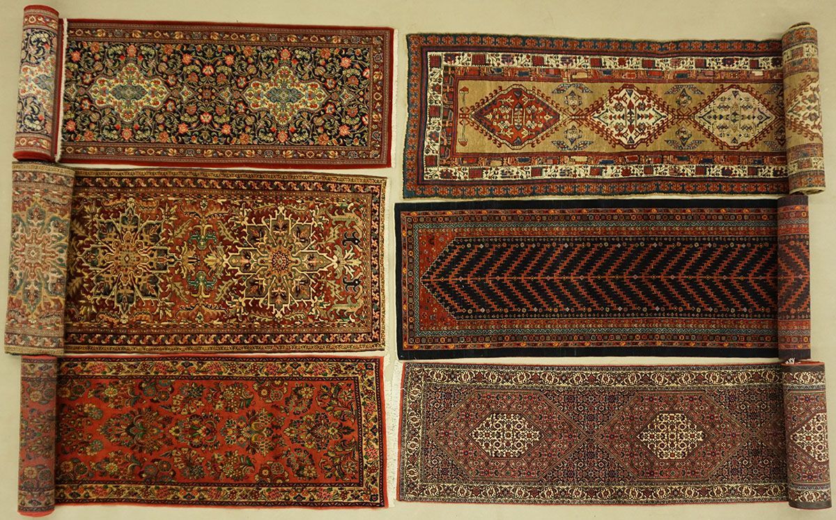Six rolled and flat Persian rugs with various patterns and colors on a neutral background