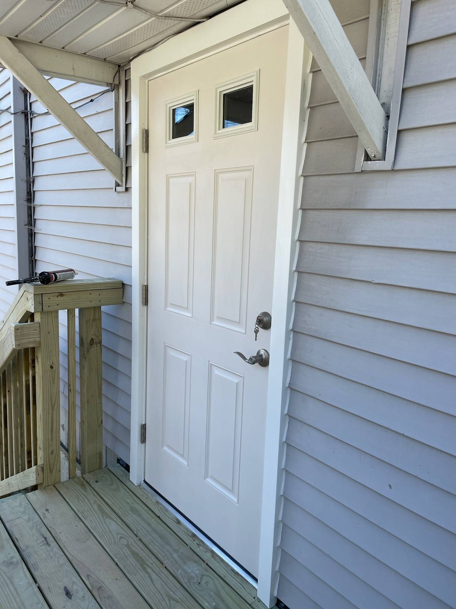 White door on a porch, light siding. Three small windows at top, gray deck and railing.