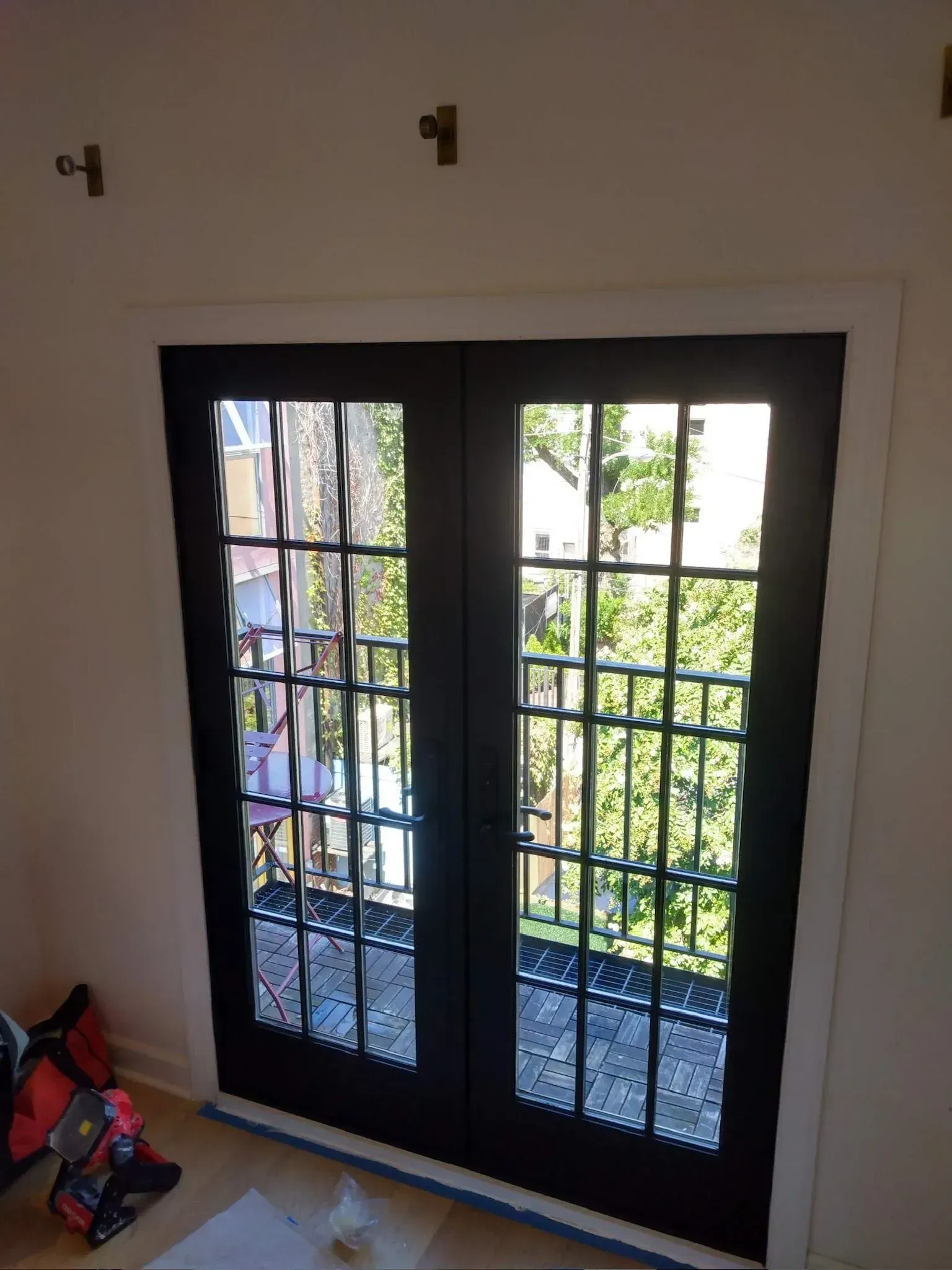 Black French doors with multiple panes, framed in white, open to a balcony with trees and buildings visible.