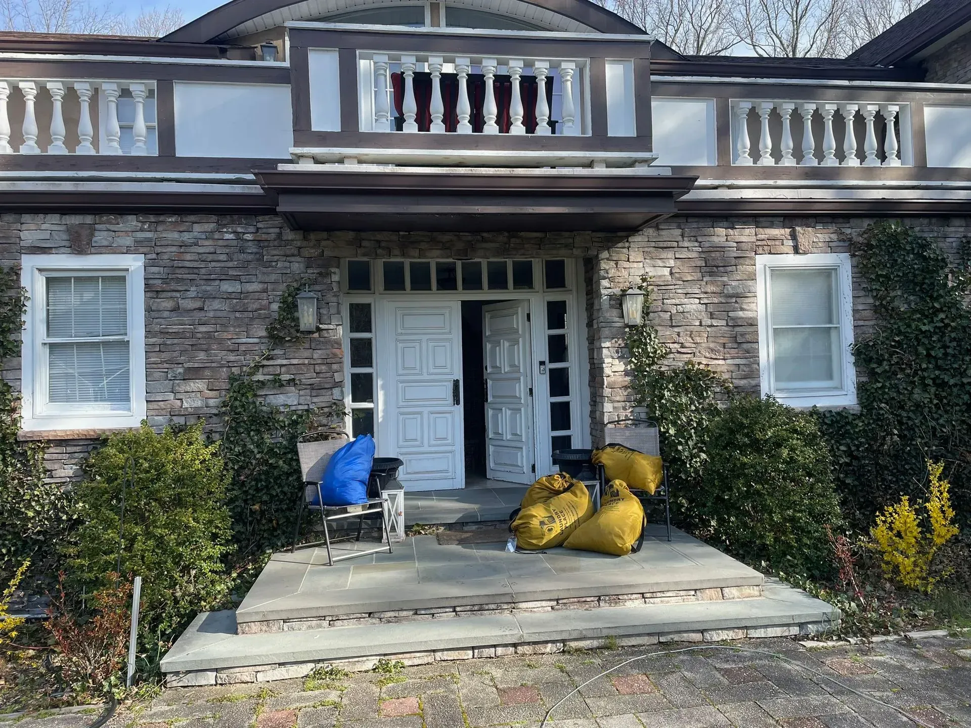 Stone house with open double doors, balcony, and porch with yard waste bags and chairs.