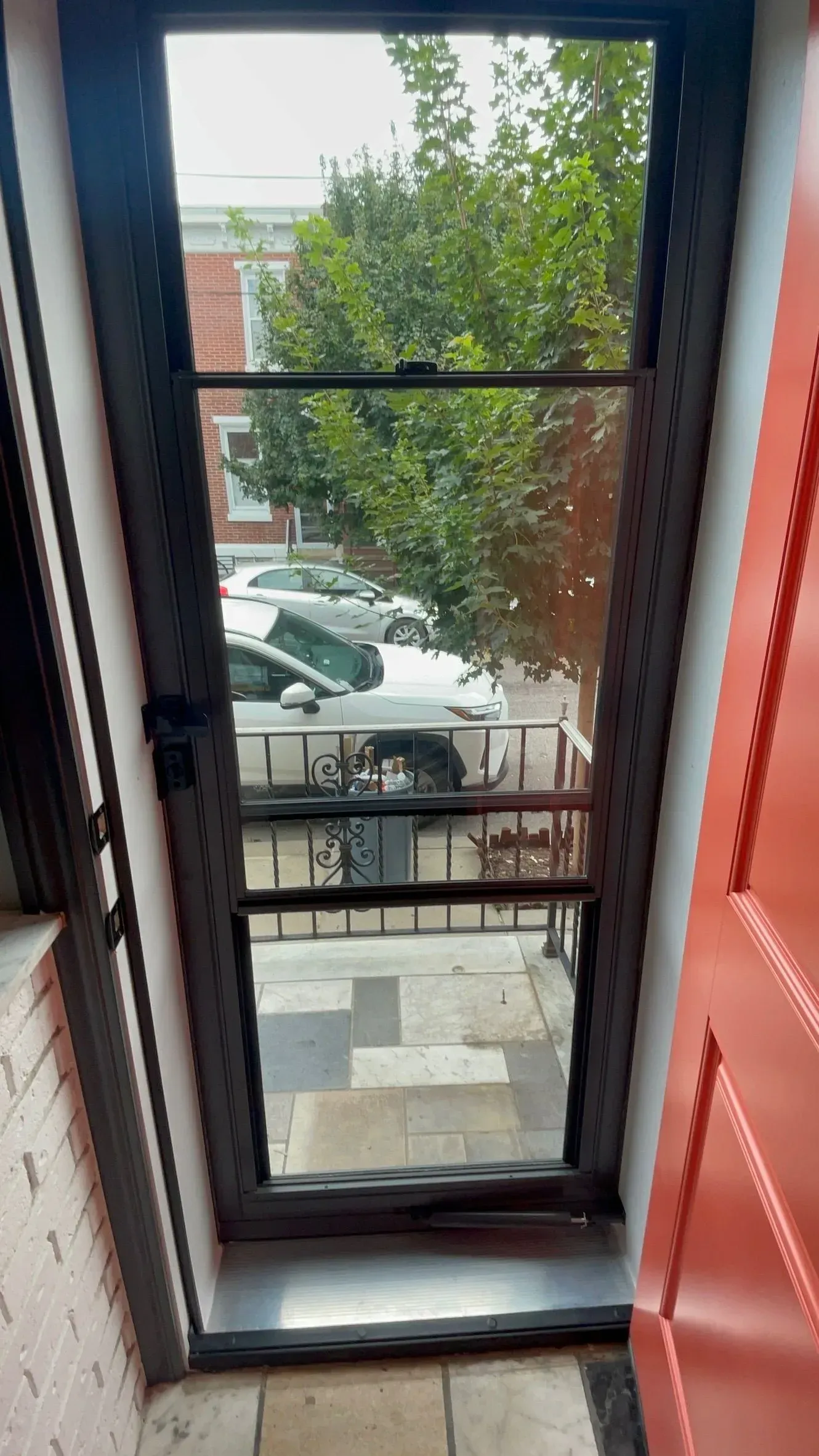 View through a dark screen door showing a street with parked cars and a tree.