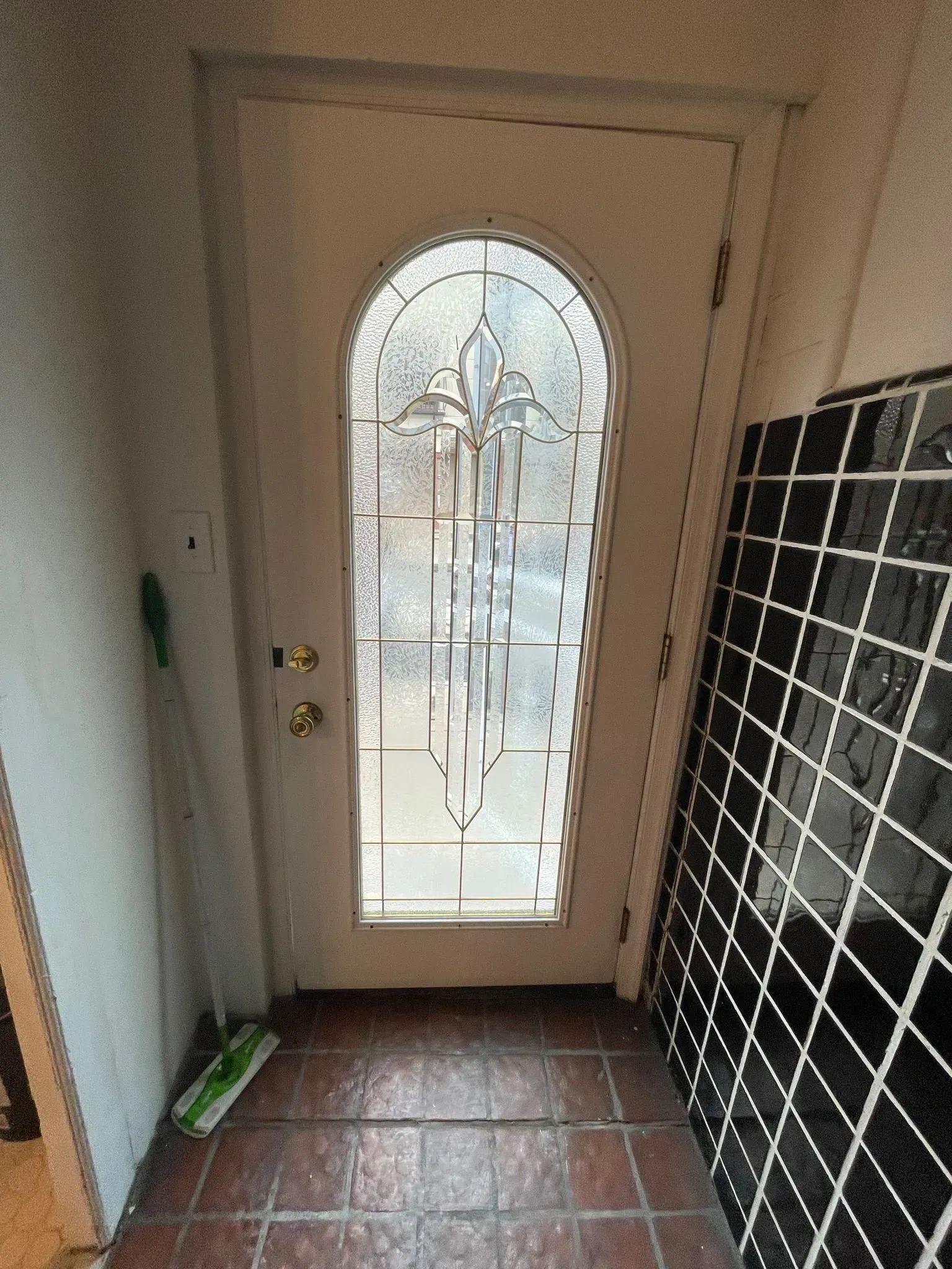 White front door with arched glass panel, small entry area with tiled floor, and some items leaning against the wall.