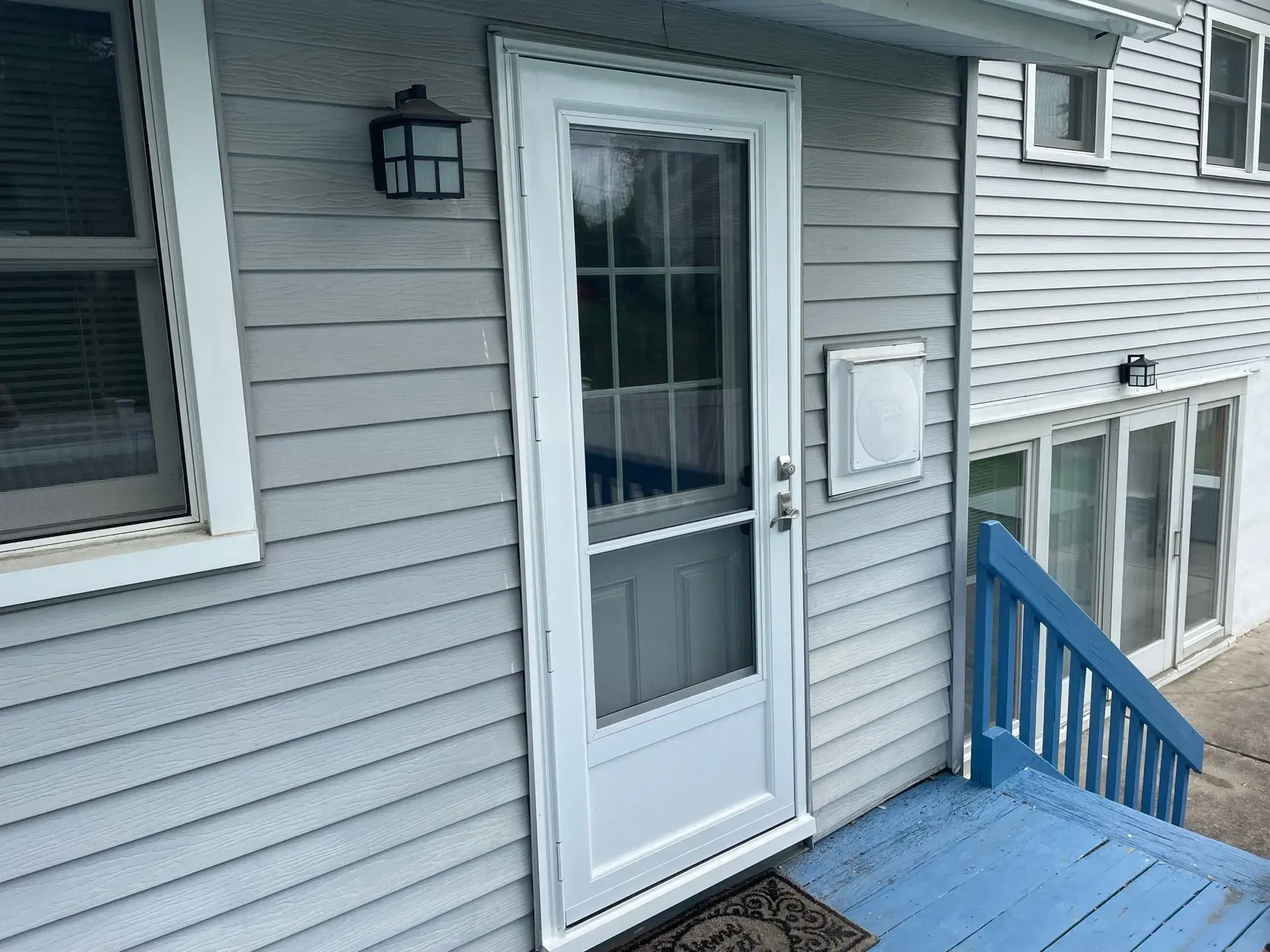 White storm door on a light blue-sided house, porch with blue stairs. A mailbox and outdoor light are visible.