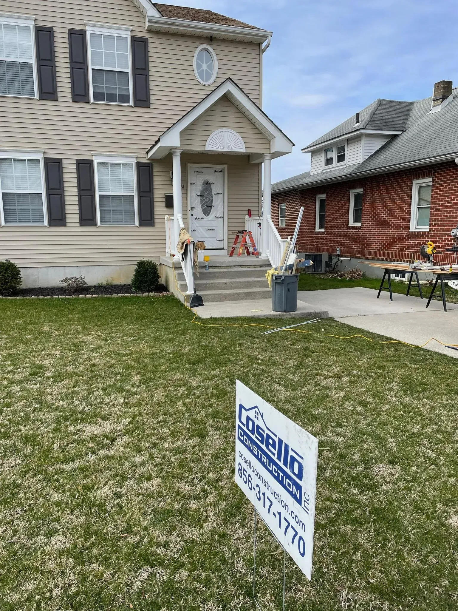 Tan house with black shutters, steps to the door, a for sale sign in front of the lawn.