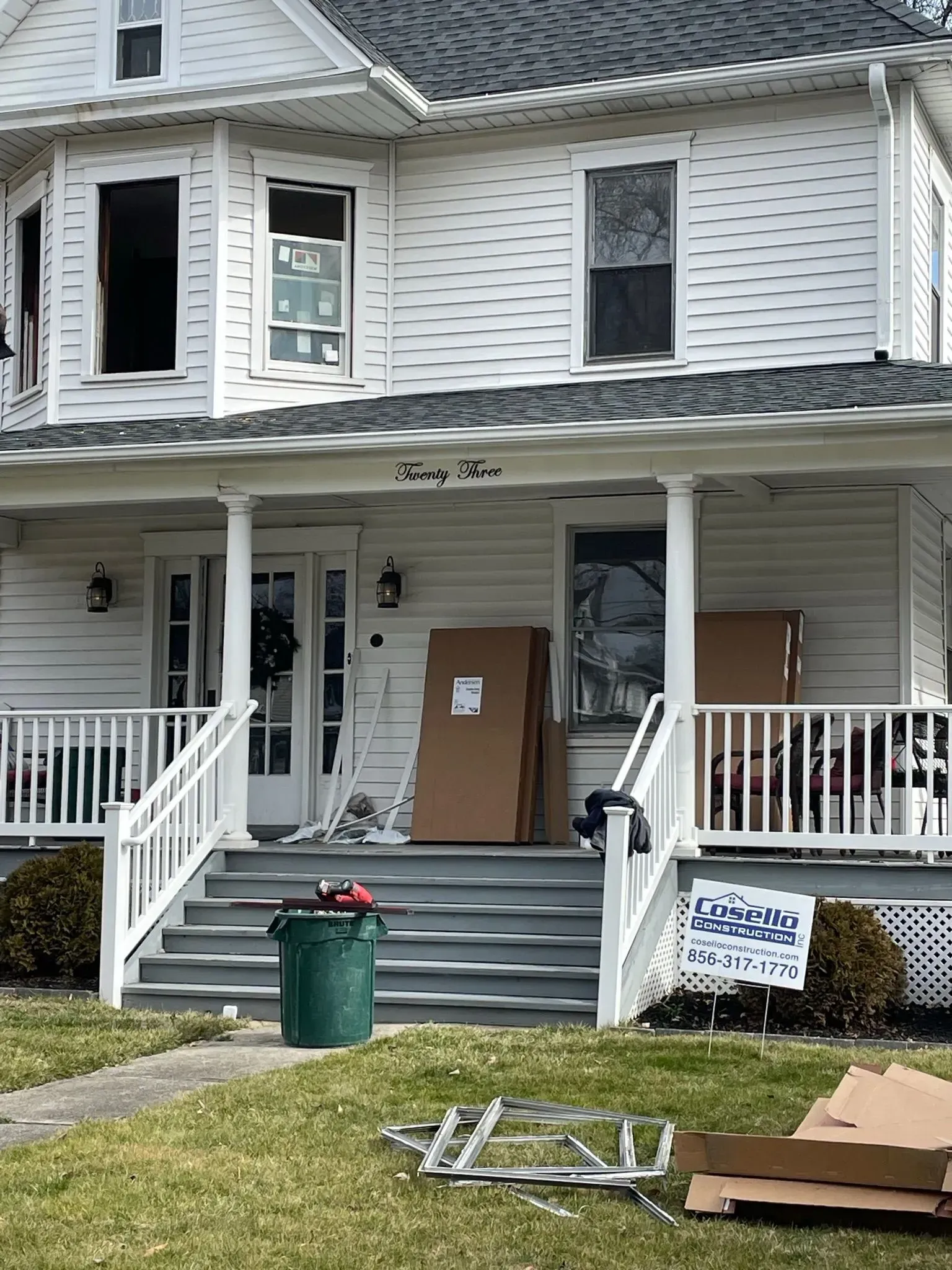 White two-story house with front porch and cardboard boxes, some windows open. Trash can and lawn debris in yard.