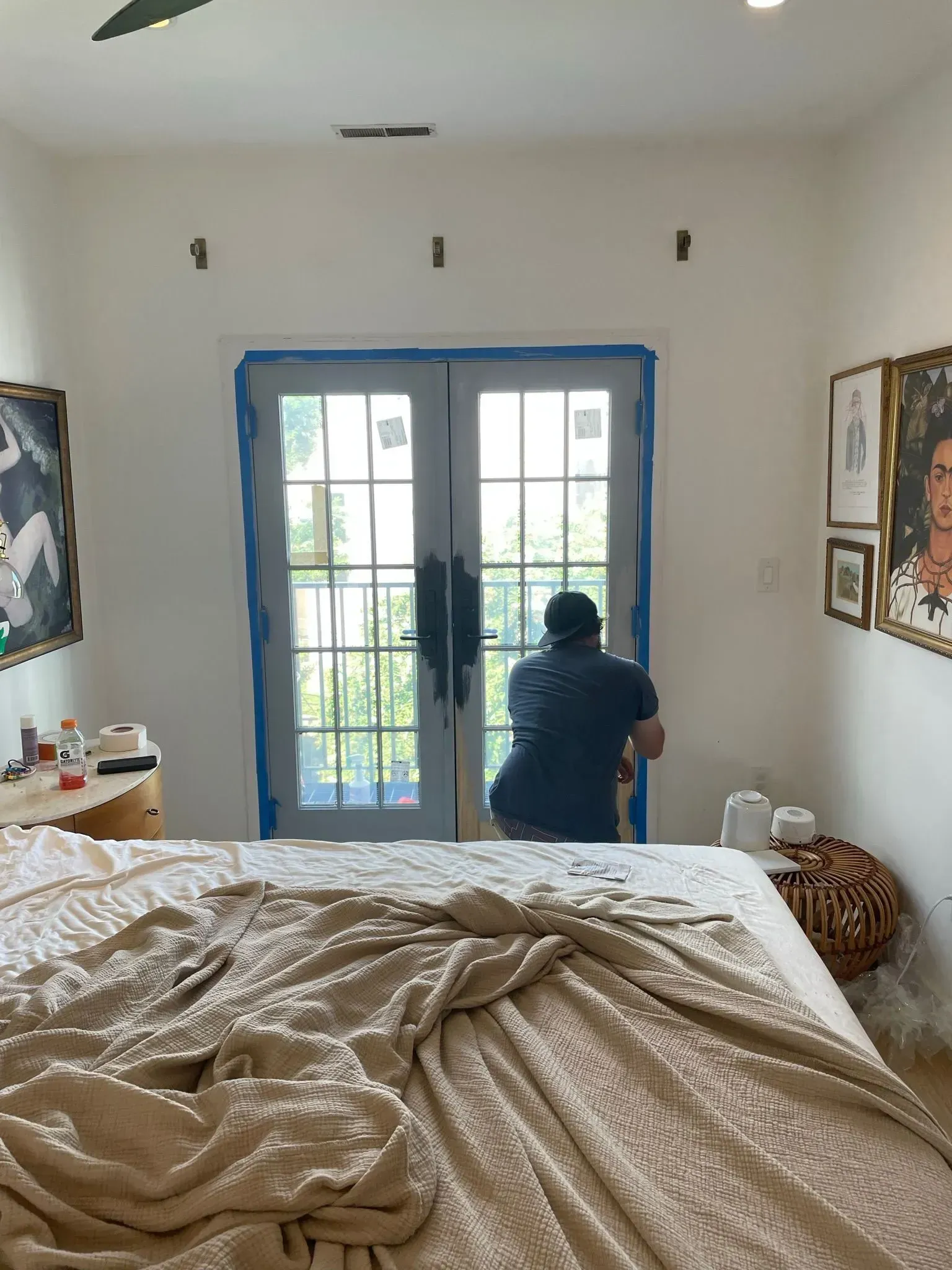 Person painting the frame of a glass door; bedroom setting. White walls, blue tape, unmade bed, art on the walls.